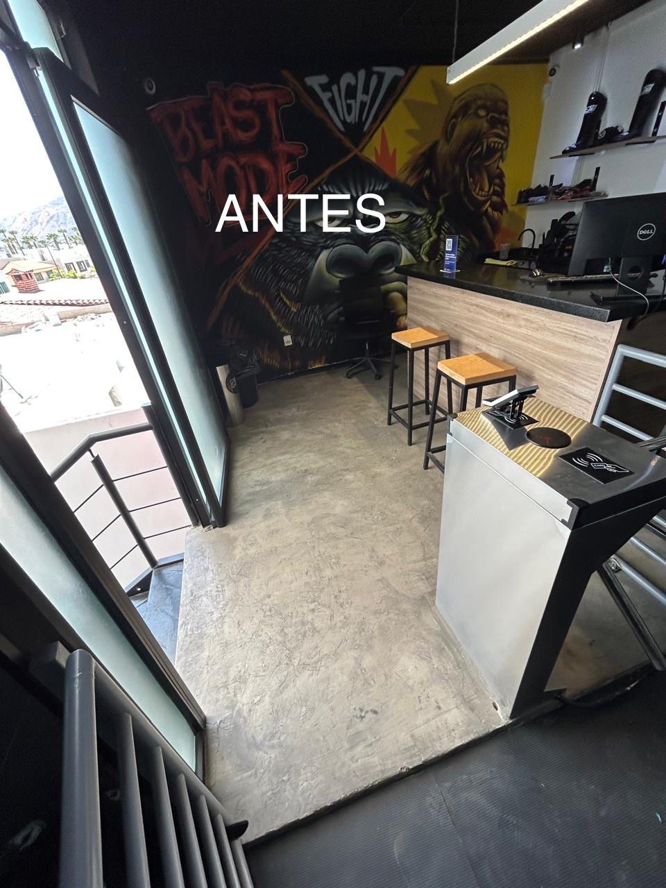 Small room with concrete flooring, bar counter with stools, computer on desk, graffiti on walls, labeled ANTES.
