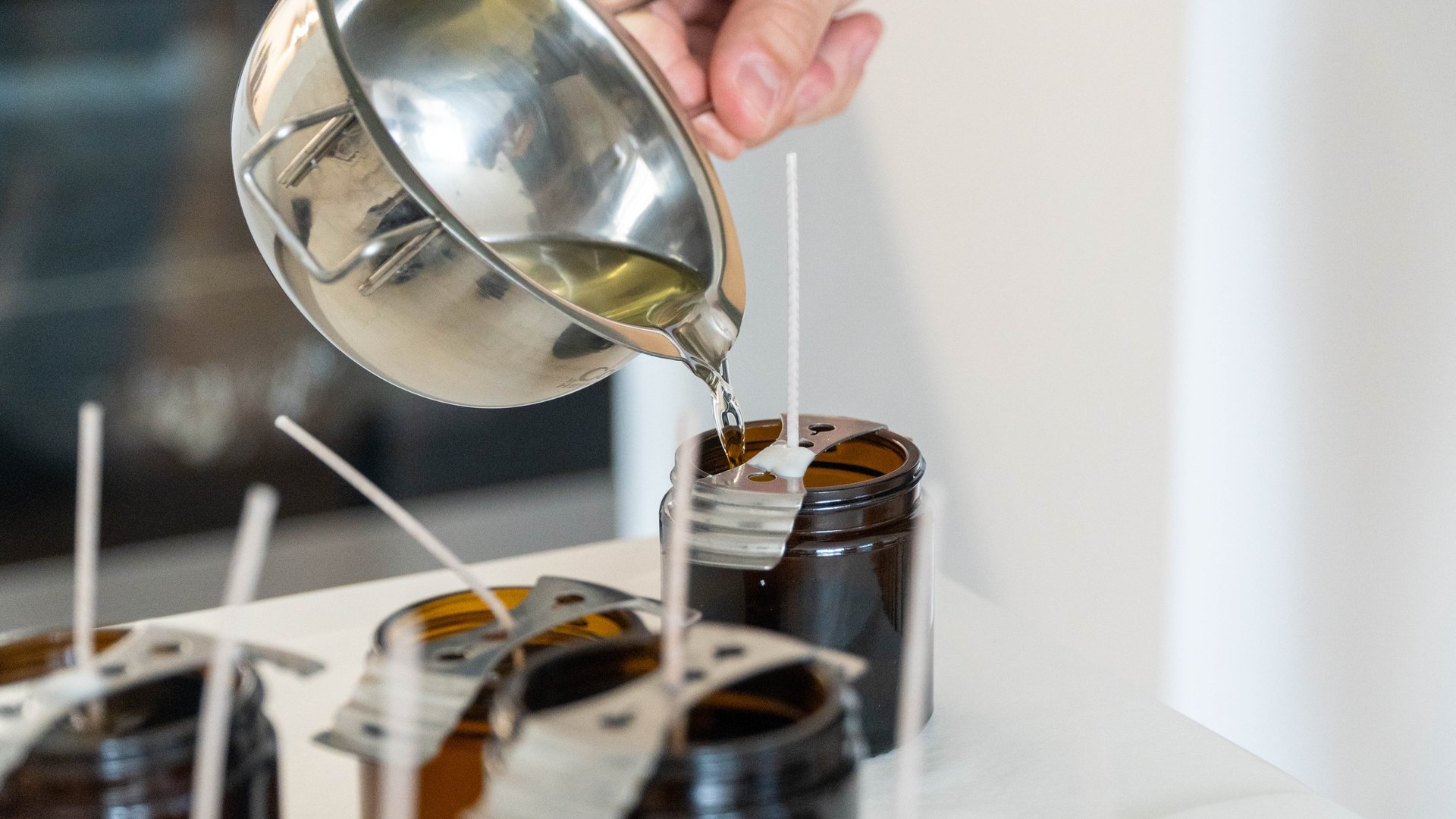Liquid wax being carefully poured into dark brown candle jars with fixed wicks. Homemade craft and artisan candle production scene.