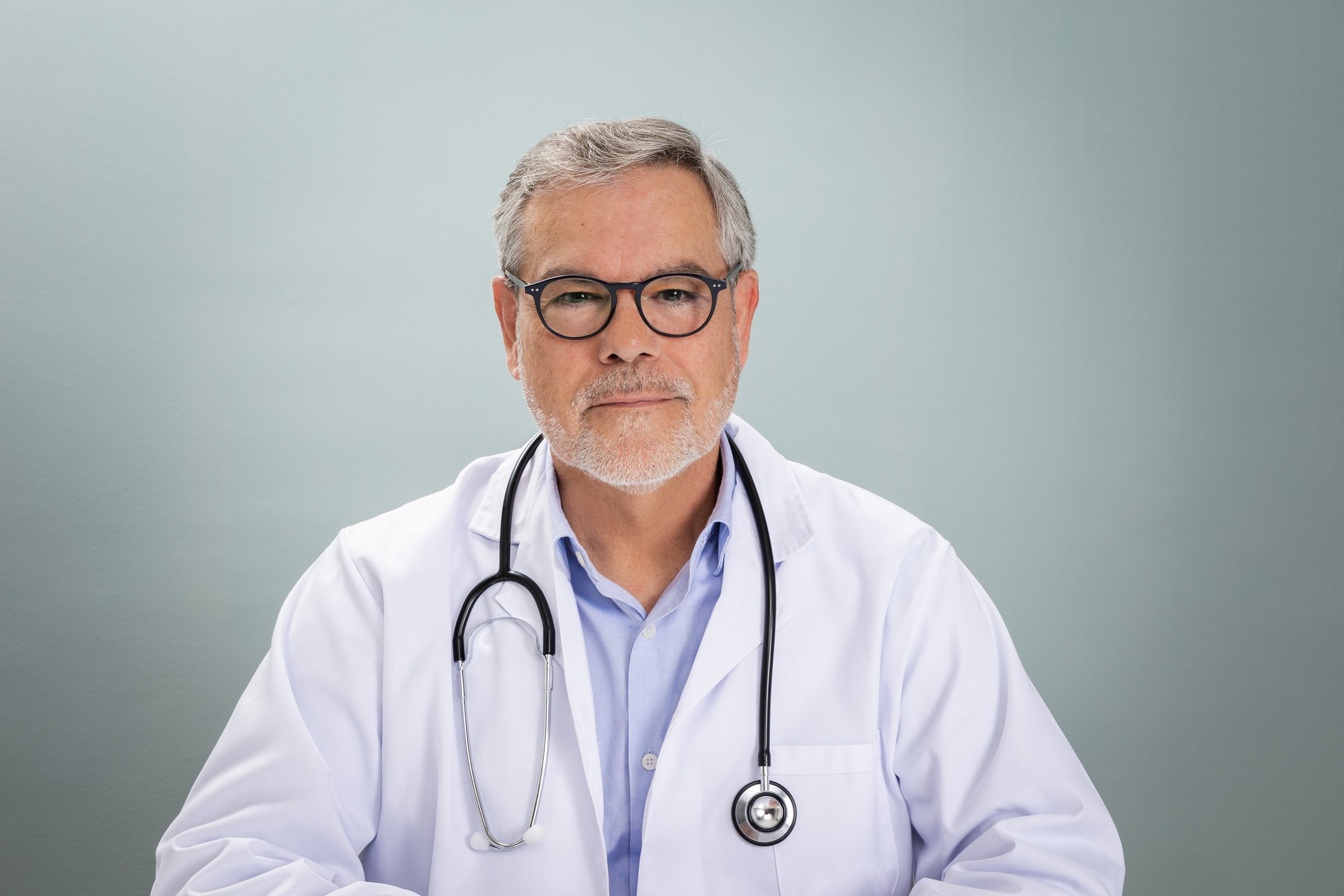An experienced doctor in a white coat sits calmly, ready for an online consultation. He wears glasses and a stethoscope, demonstrating professionalism and trust.