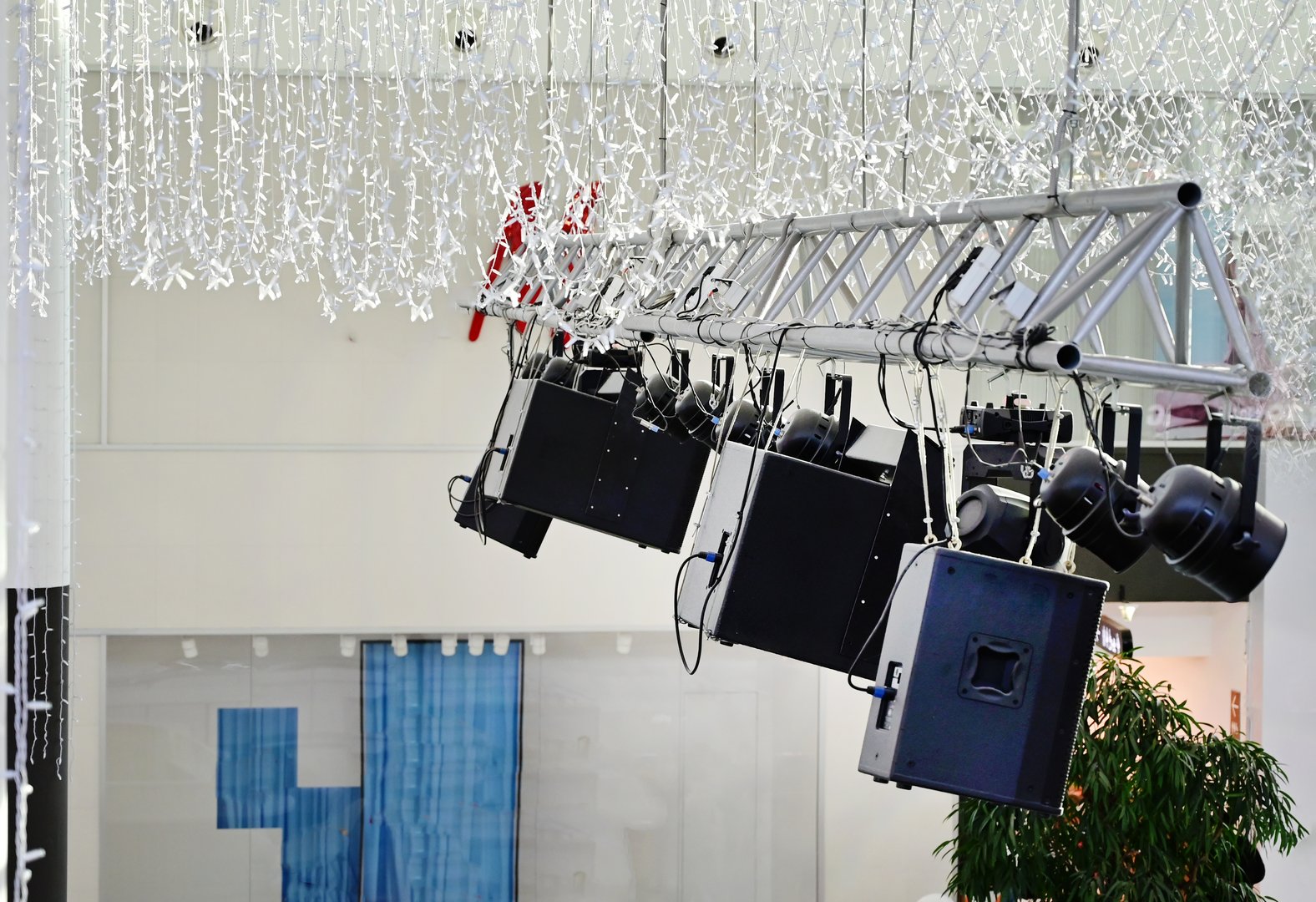 Suspended audio equipment hangs from a truss overhead in a bright indoor space, preparing for an upcoming event with decorative lighting in view.
