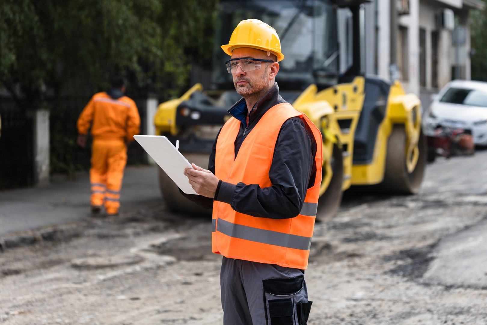 A construction supervisor in a reflective vest checks plans on a clipboard while monitoring roadwork in a city setting, with machinery active in the background.