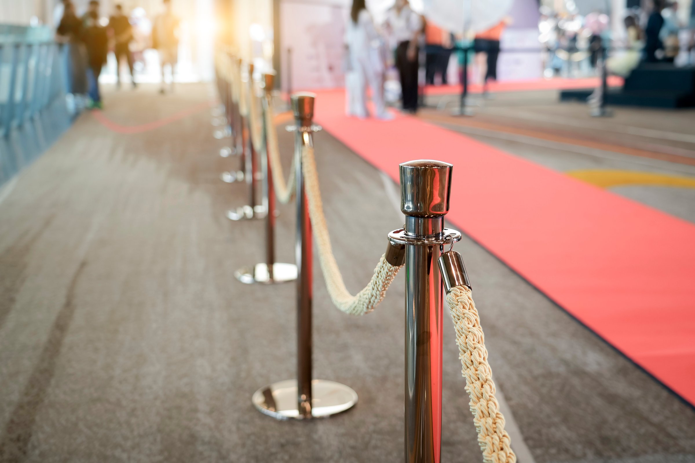 A long row of polished stanchions and braided ropes border a red carpet. People walk by in the background at what appears to be some type of event.