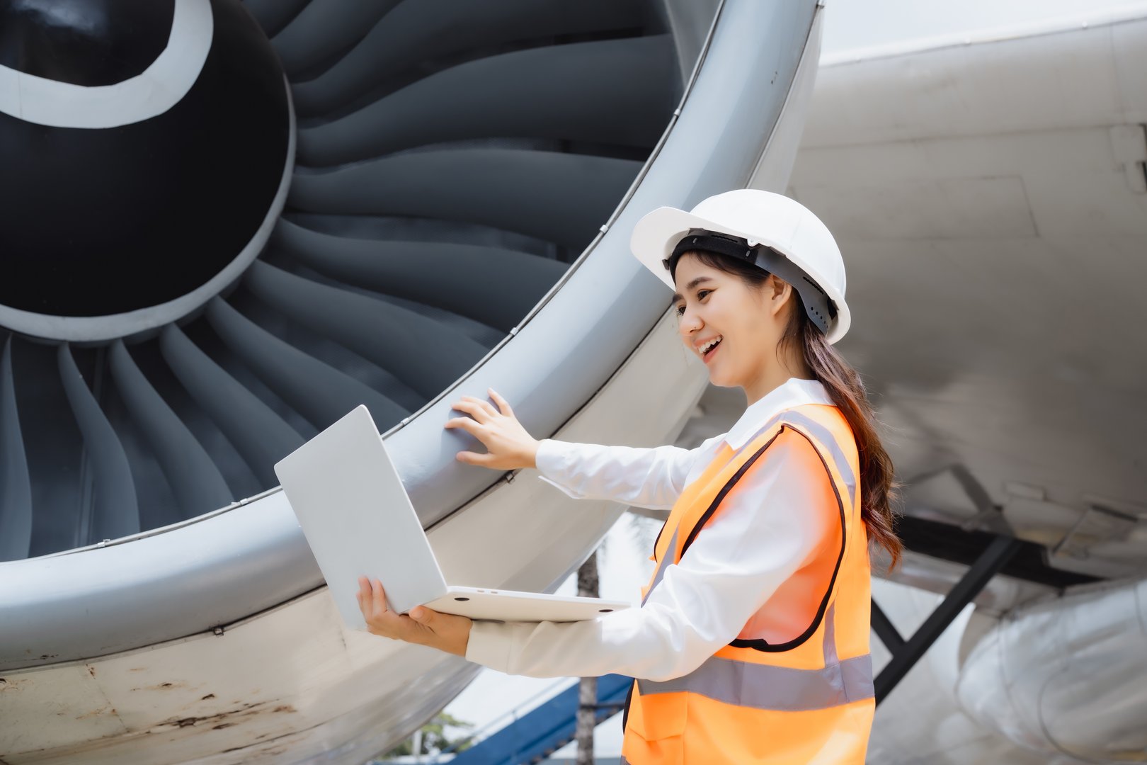Asian woman aviation engineer wearing safety helmet and reflective vest holding tablet in hangar