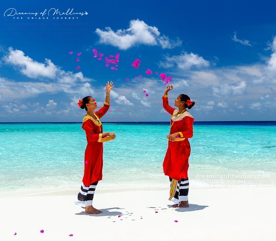 Two women in red dresses toss pink flowers on a beach with turquoise waters and blue sky in the Maldives.
