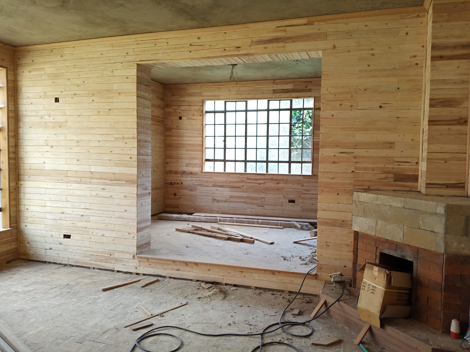 Interior under renovation with wooden wall paneling, a brick fireplace, parquet flooring, and construction materials.