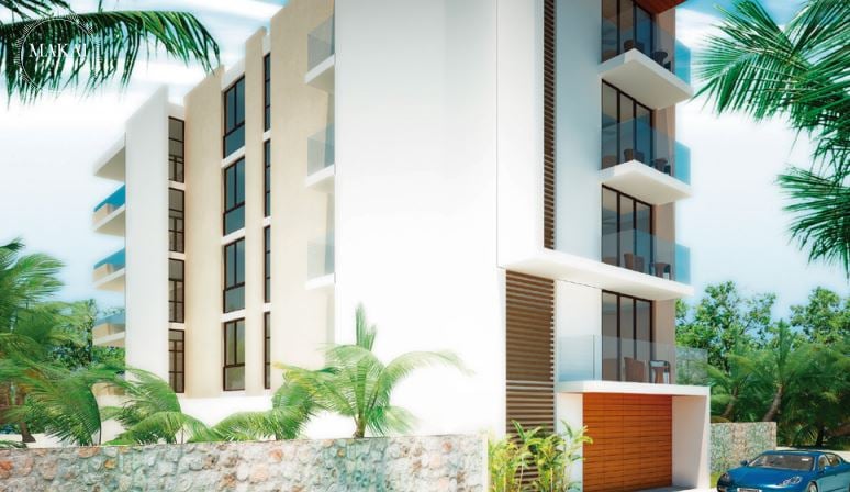 Modern multi-story building with large windows and balconies, surrounded by palm trees and a stone wall.