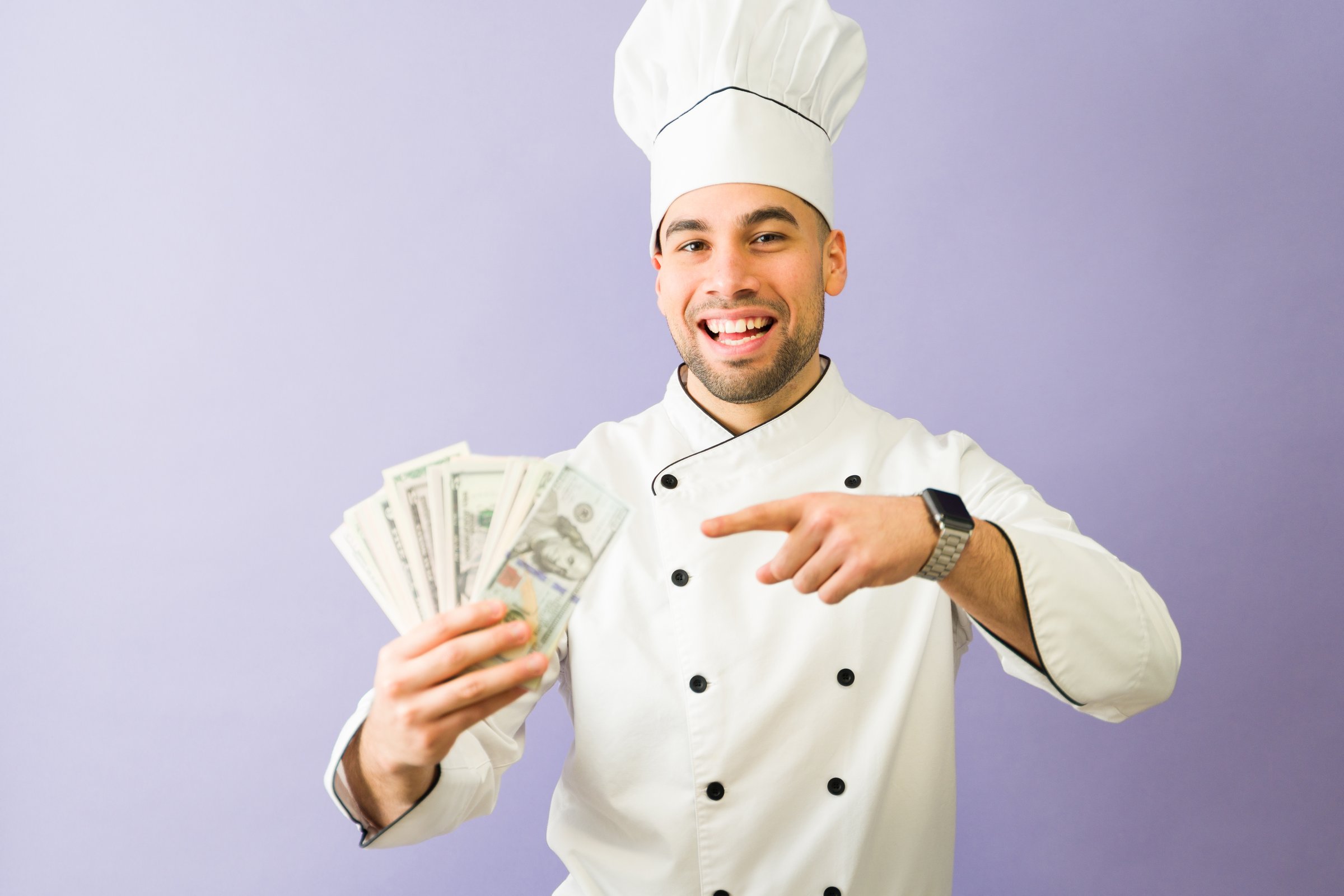 Proud latin chef pointing at dollar bills smiling at camera wearing white uniform and chef hat over purple background