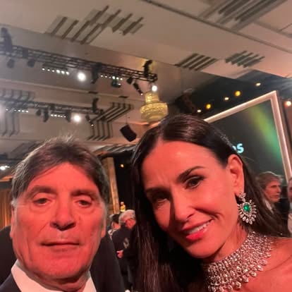 A man and a woman in formal attire at a glamorous event with chandeliers and stage lights in the background.