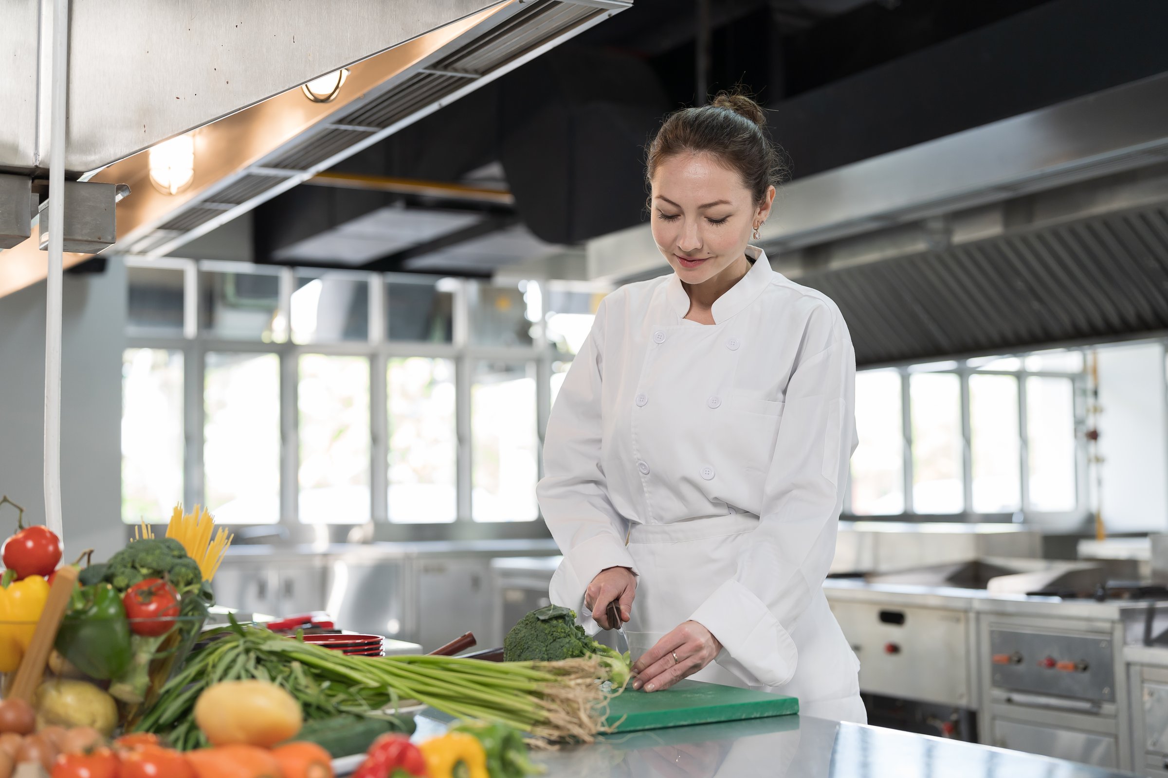 Restaurant kitchen. Female chef preparing meal in kitchen, cutting board with vegetables, wearing white apron. Female cooks preparing food in kitchen at restaurant