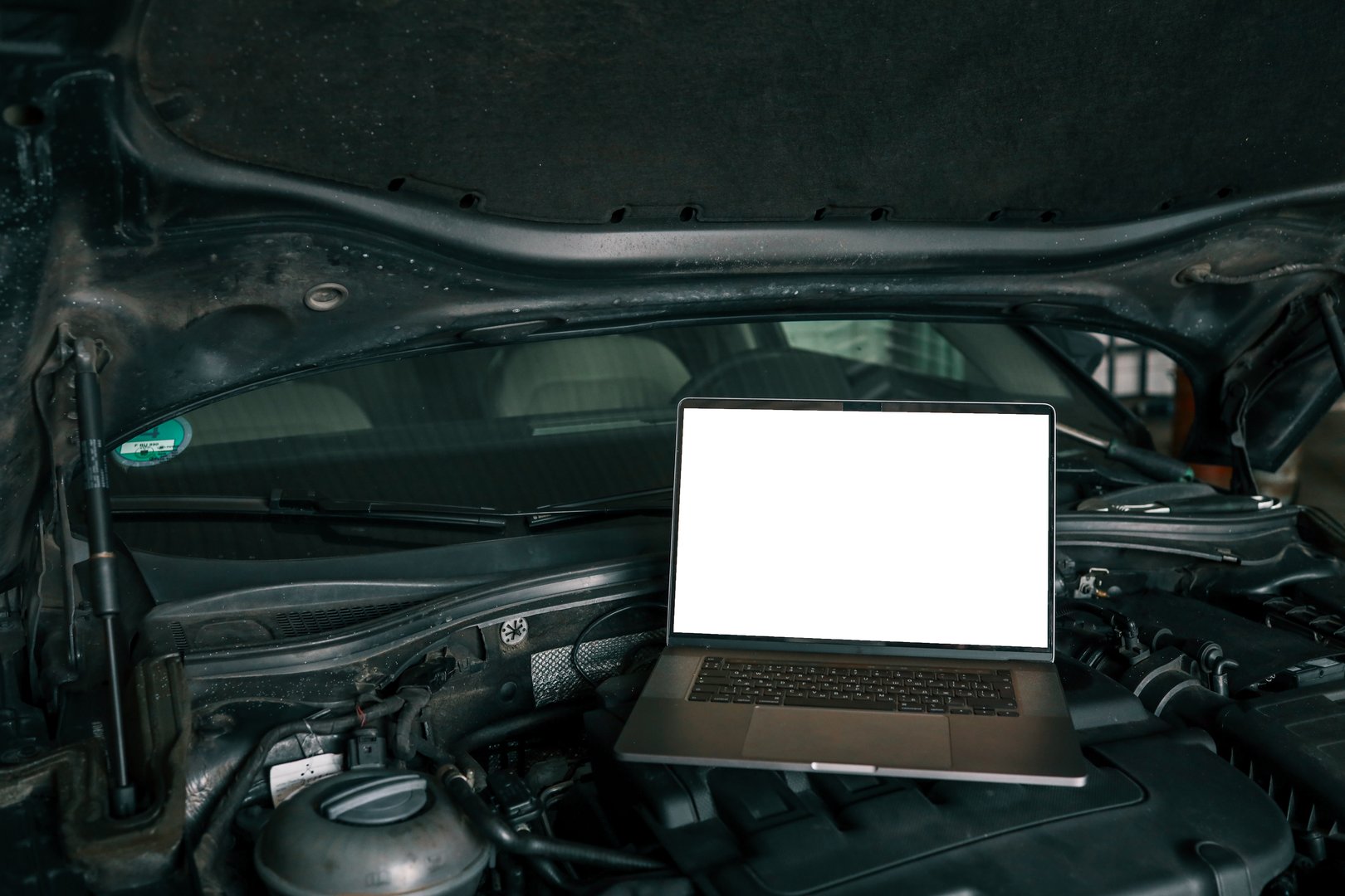 Laptop placed on a car engine under an open hood in a garage. Represents modern automotive diagnostics and maintenance using computer technology.