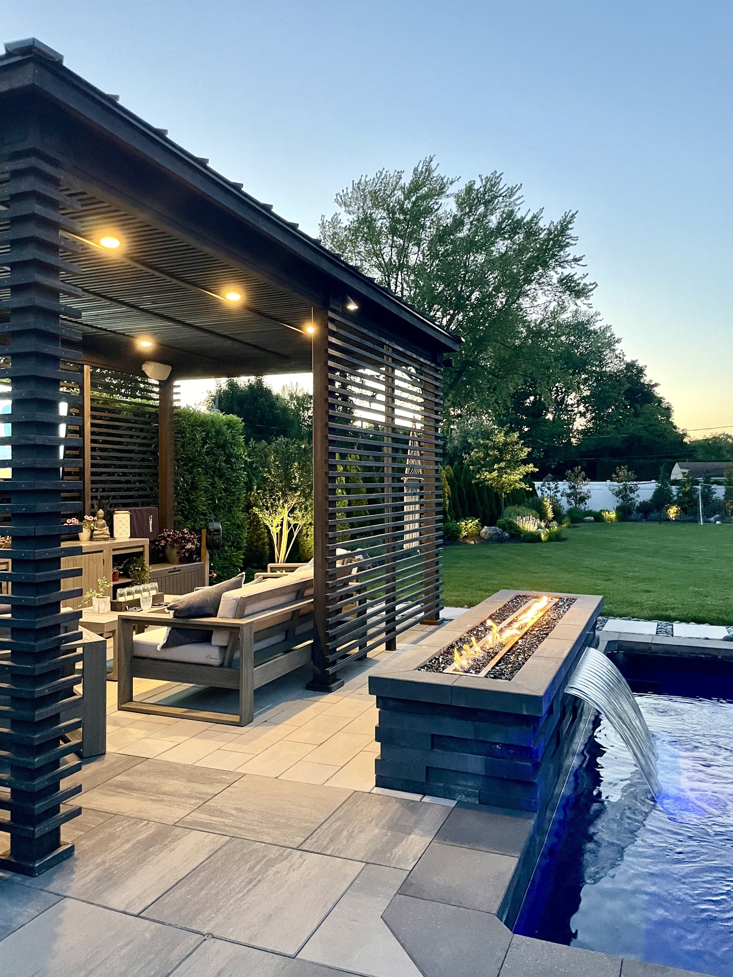 High-end outdoor living space with backyard patio, pool with waterfall, and stunning entertainment space surrounded by lush gardens, flowers, shrubs and tropical trees.