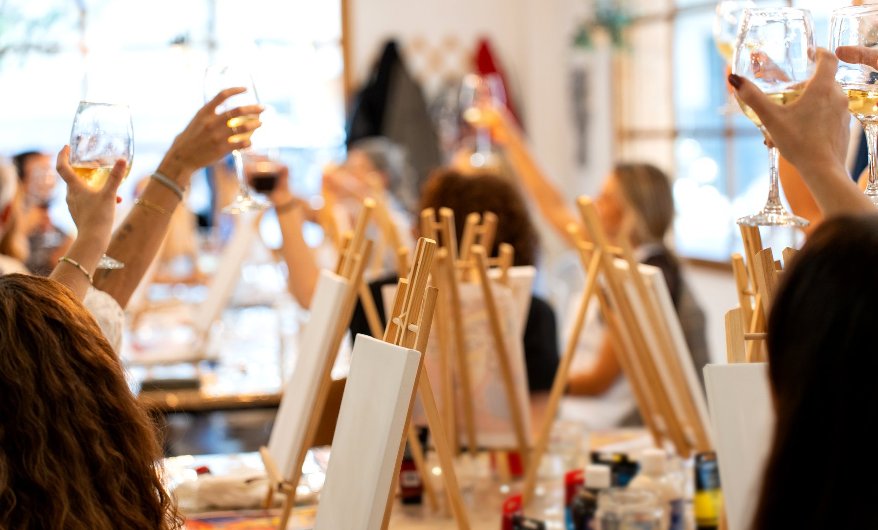 Women lifting wine glasses, celebrating and creating art together during a social painting class, fostering creativity and fun