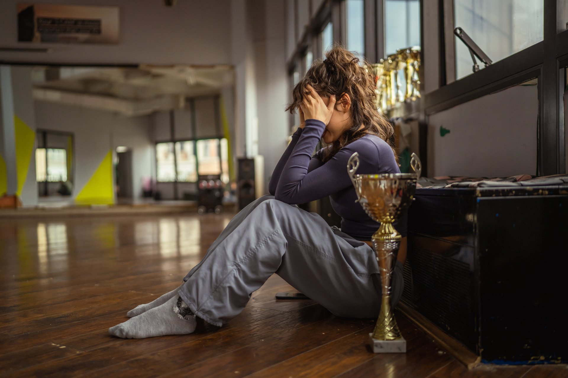 Young woman dancer sitting on wooden floor in a studio, experiencing burnout and stress, feeling overwhelmed and saddened by pressure to perform, next to trophy