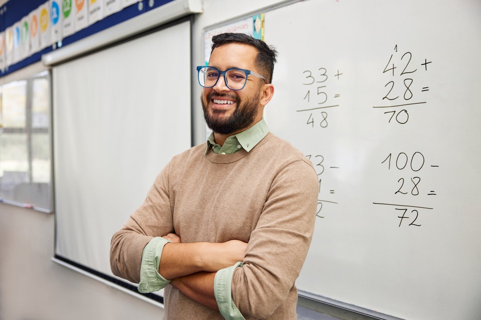 Happy teacher explaining mathematics at whiteboard in classroom. Portrait of smiling mathematics tutor explaining addition concept to children in classroom. Successful teacher standing in front of whiteboard with crossed arms after explaining addition to students.