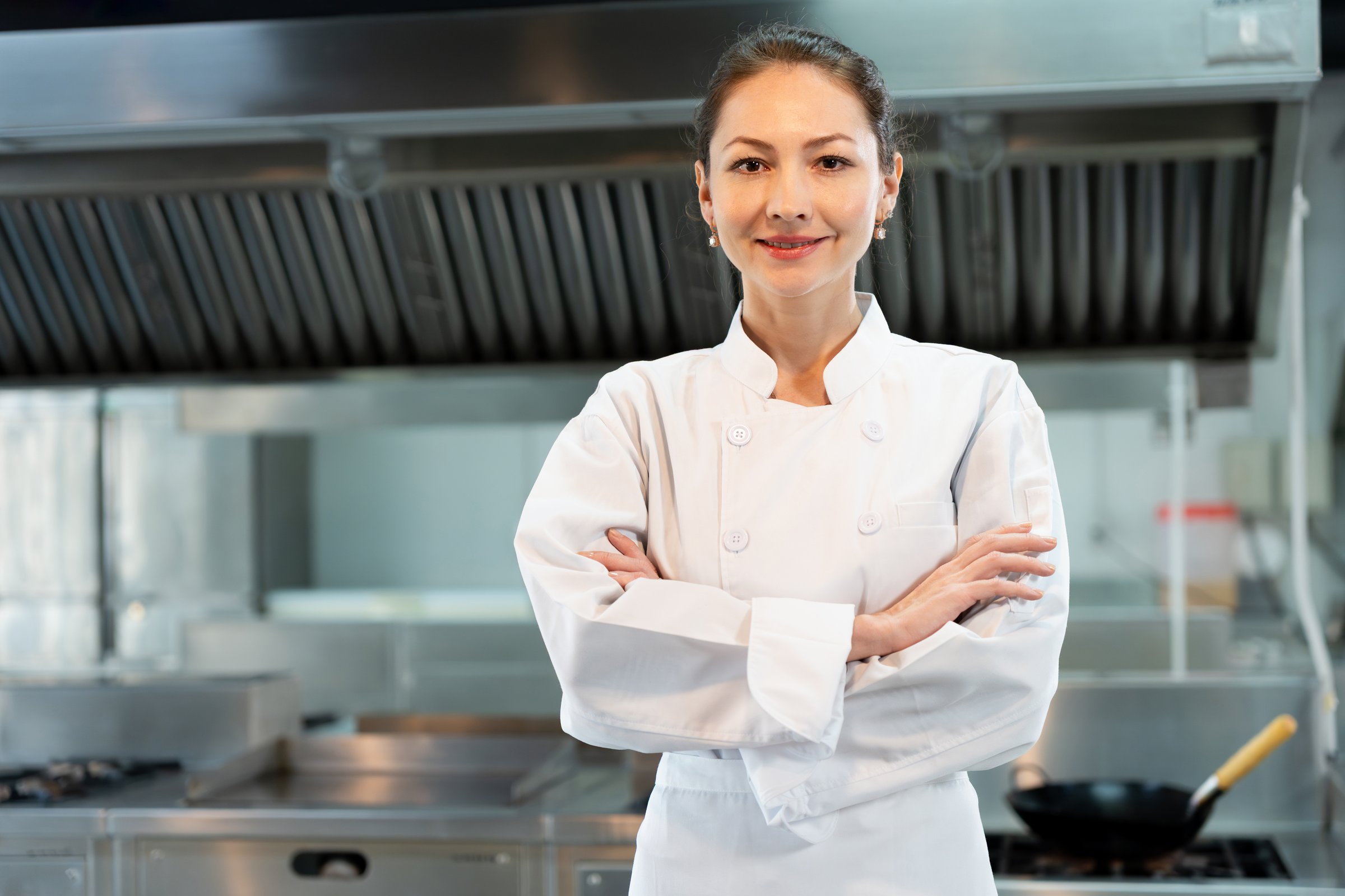 Confident Female Chef Posing in a Professional Kitchen, Portrait of a Smiling Woman Chef in White Uniform, Experienced Culinary Professional Stands with Arms Crossed in a Commercial Kitchen