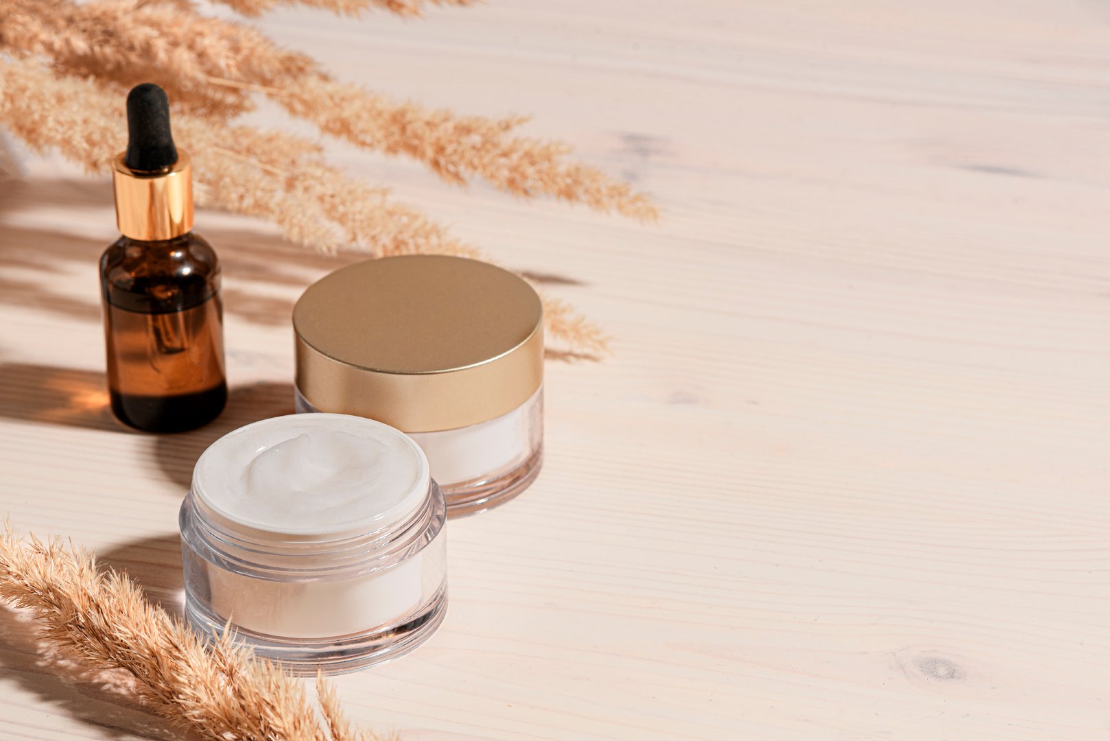 Cosmetic serum and beauty cream products with dry reeds or pampas grass on wooden background, trendycomposition with hard light. Facial anti age natural skin care, copy space.