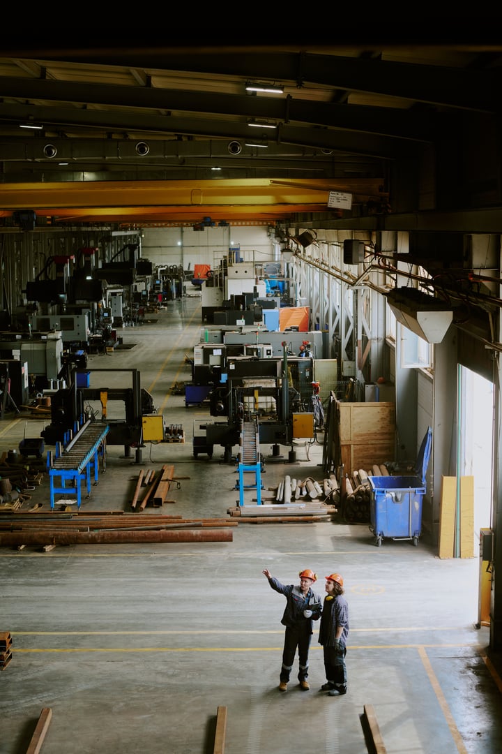 Two middle aged Caucasian women wearing safety helmets standing in large industrial factory, discussing machinery layout and pointing toward equipment on production floor