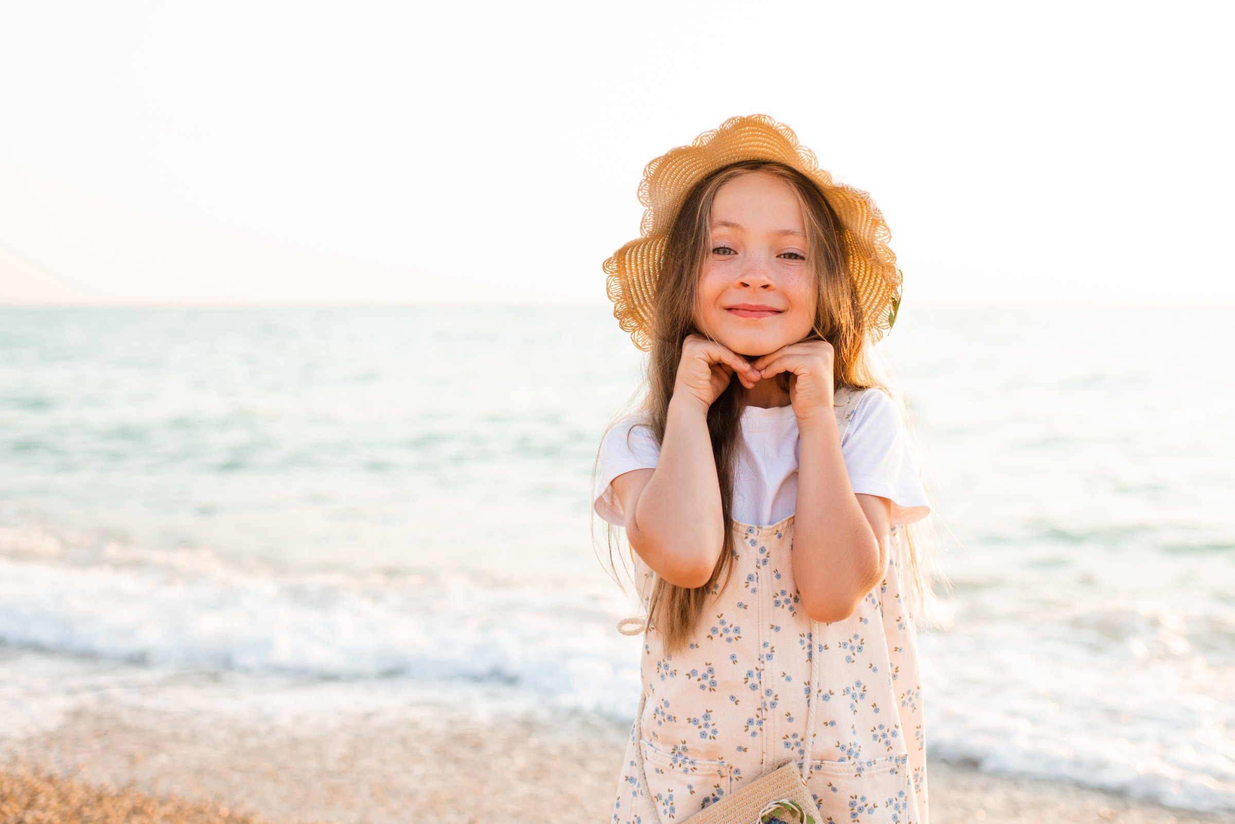 Smiling baby girl wearing straw hat and denim dress standing at beach