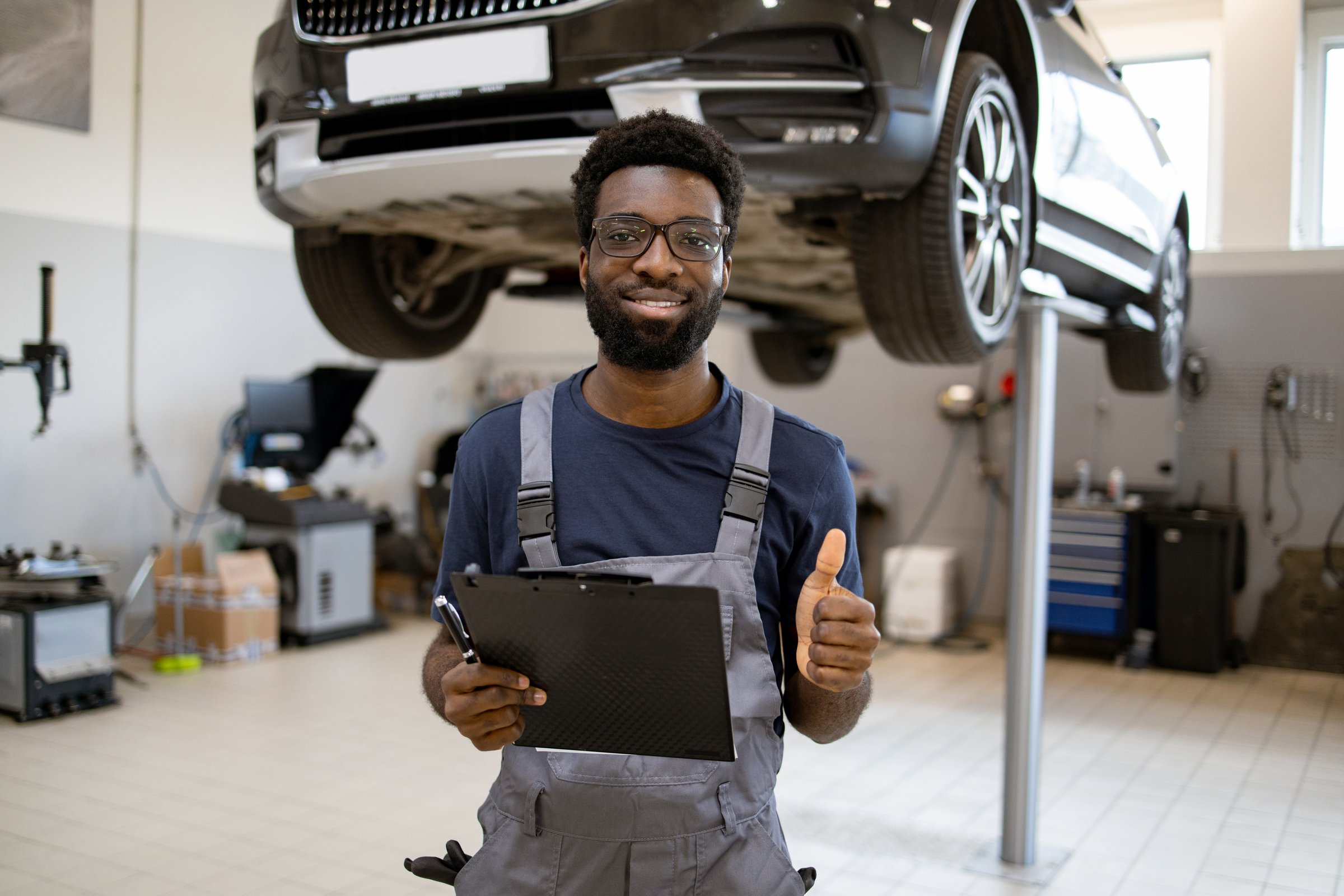 Confident auto mechanic smiling with clipboard and thumbs up in repair workshop. Professional technician in uniform standing in front of car on lift, displaying approval with positive gesture