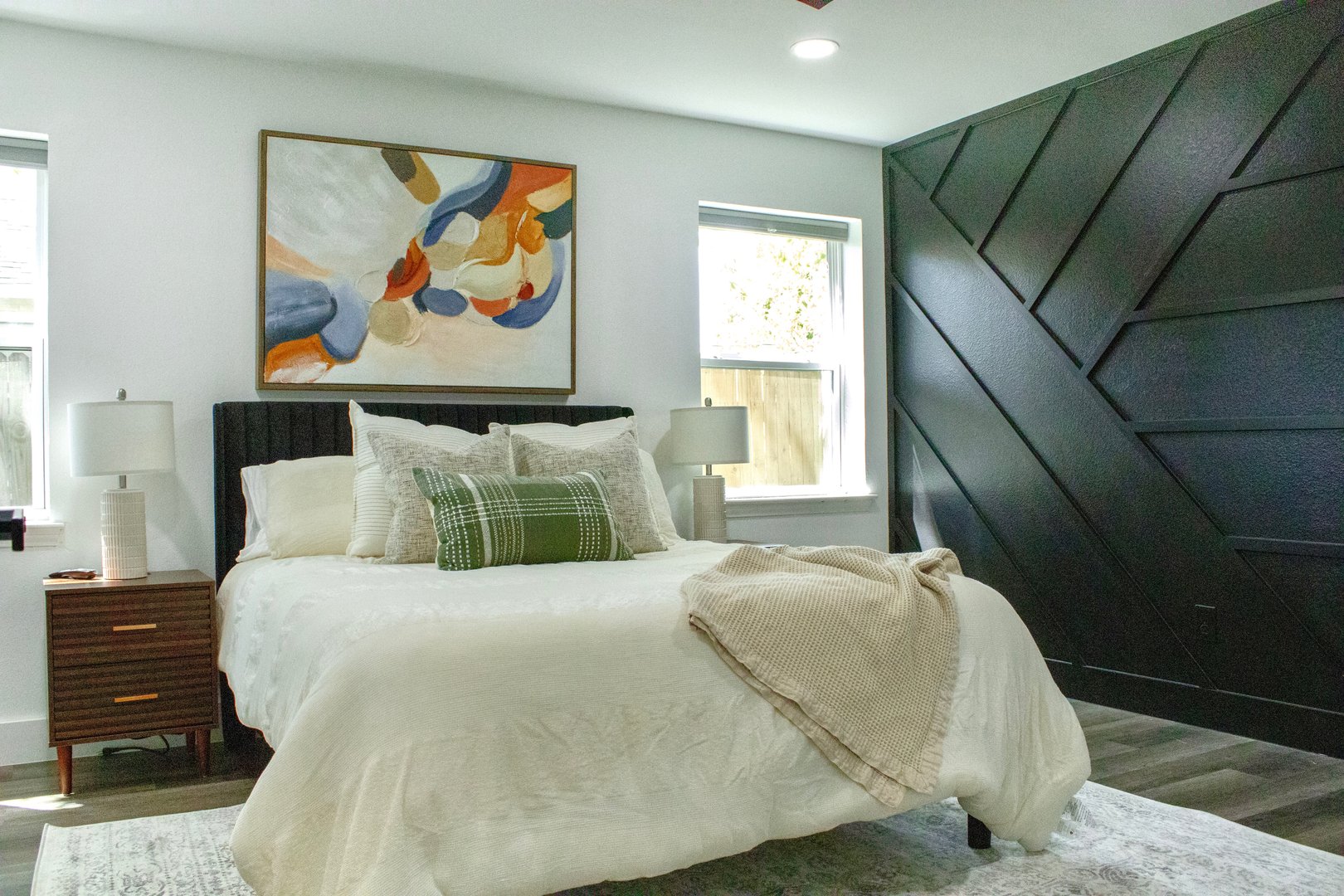 Modern bedroom with abstract art, dark accent wall, and two bedside tables with lamps. Bed with white linens and green pillows.