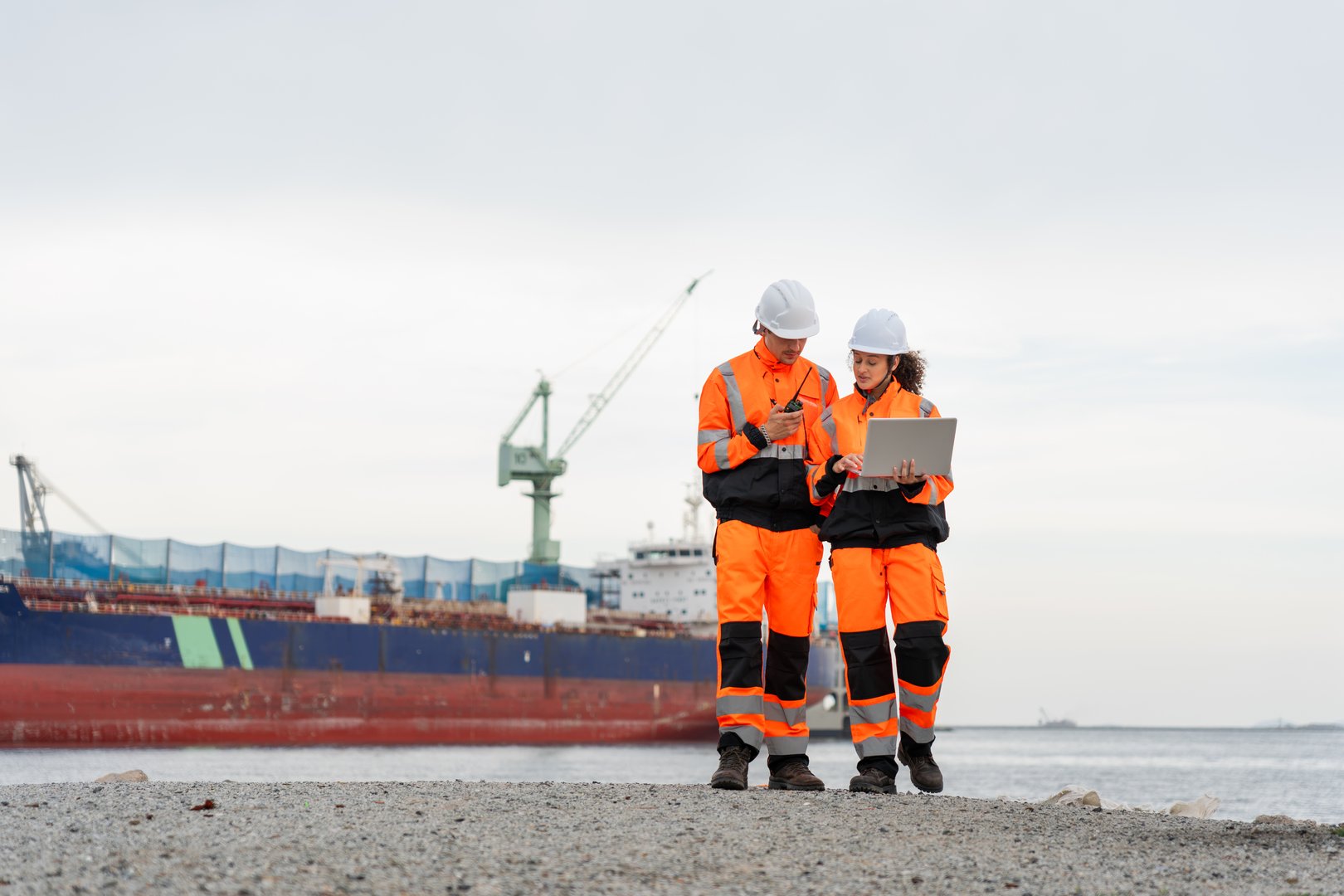 Industrial Engineers Collaborating at Harbor Site, Dock Worker in Safety Uniforms Using Technology for Port Operations, Maritime Safety Engineers Working at Port Terminal with Laptop and Walkie Talkie