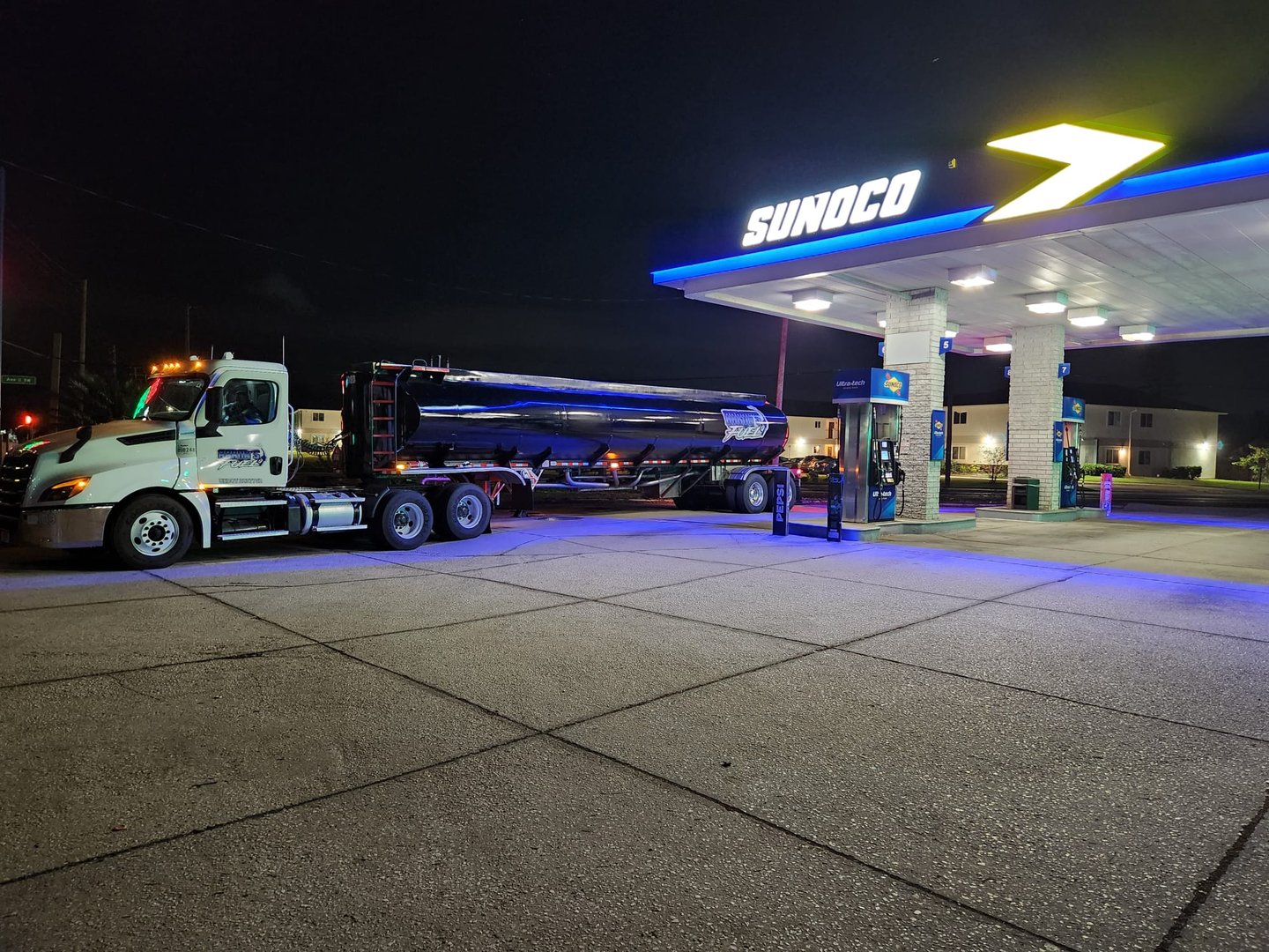 A fuel truck parked at a Sunoco gas station at night, illuminated by station lights, with a tanker attached.
