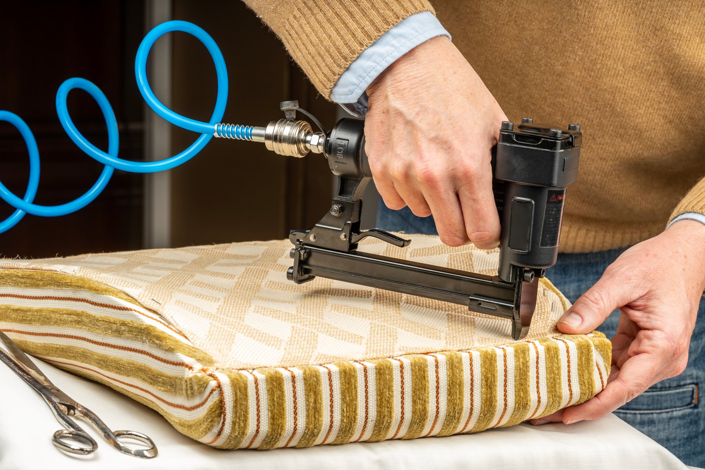 Close-up of an upholsterer's hands stapling fabric on a chair seat with a pneumatic stapler.