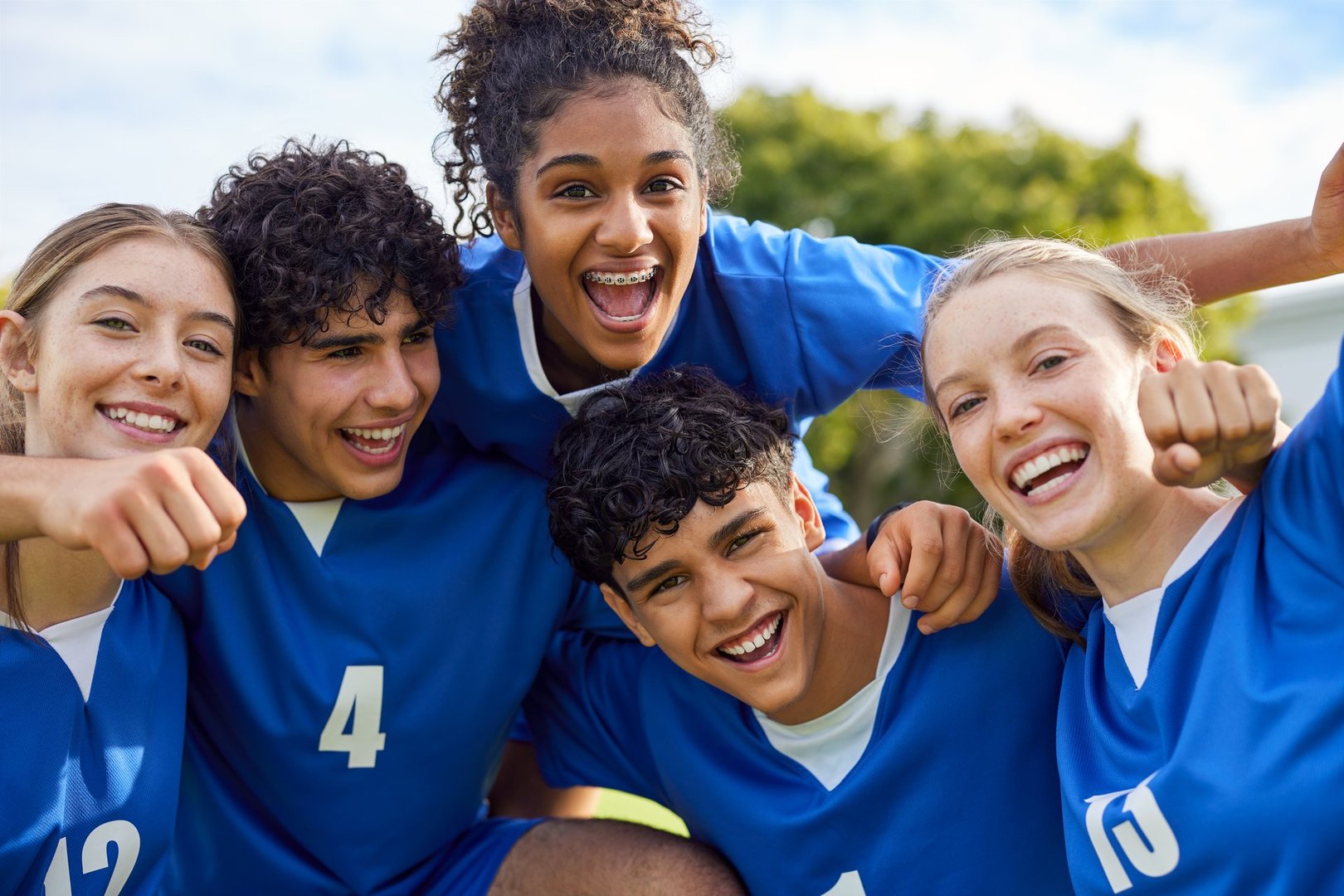 Smiling group of young teammates celebrating a joyful win of football match. Portrait of sports players of guys and girls cheering together after match. Excited young sports team of guys and girls posing with energy and happiness on the field after a soccer goal.
