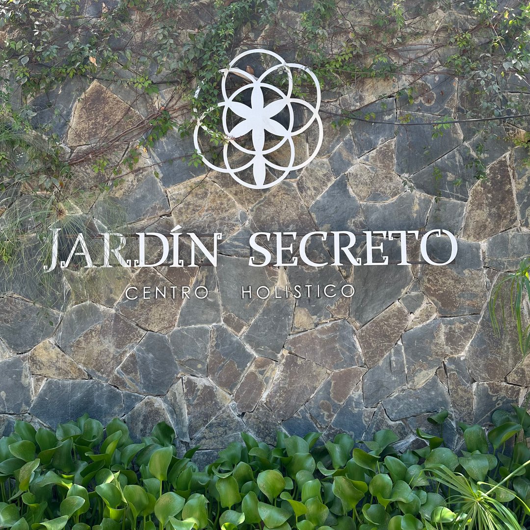 Stone wall with "Jardín Secreto Centro Holístico" and a floral symbol, surrounded by greenery.