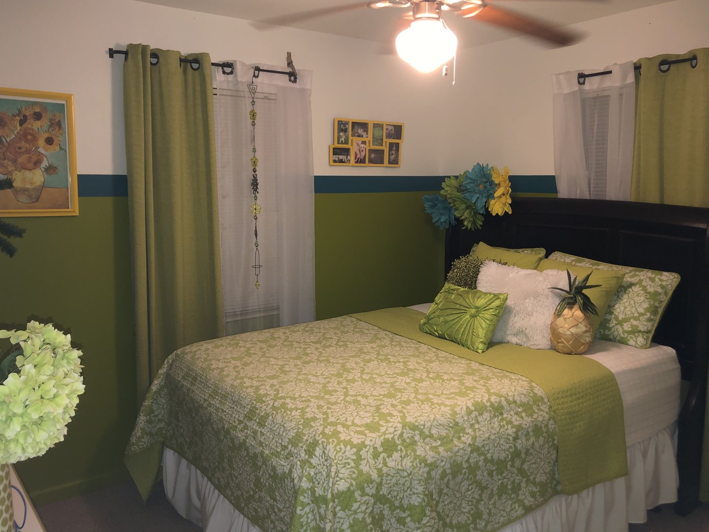 Cozy bedroom with green decor, floral bedspread, and decorative pillows. Green curtains and sunflower artwork on wall.
