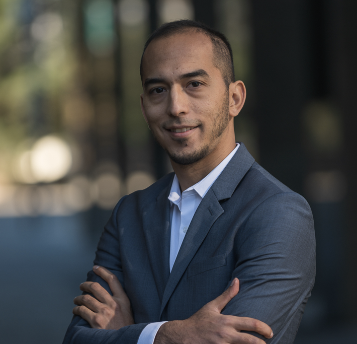 Man in a gray suit stands outdoors with arms crossed, smiling slightly against a blurred background.