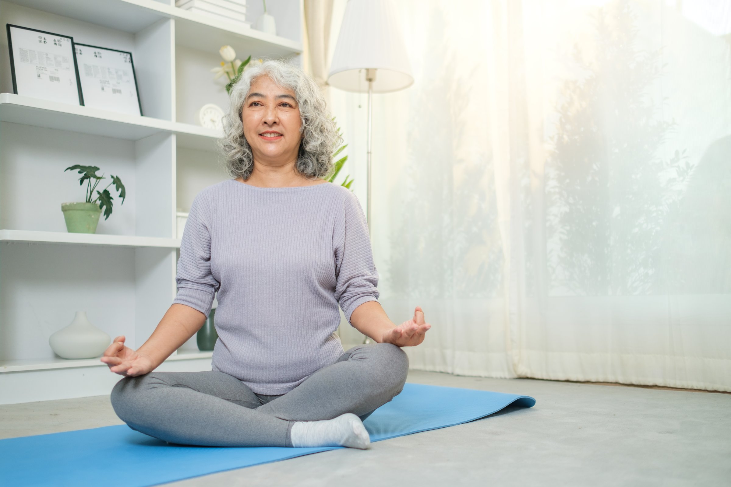 Calm mature woman doing breath exercises during yoga session on mat in the living room. Retirement and healthy lifestyle concept.