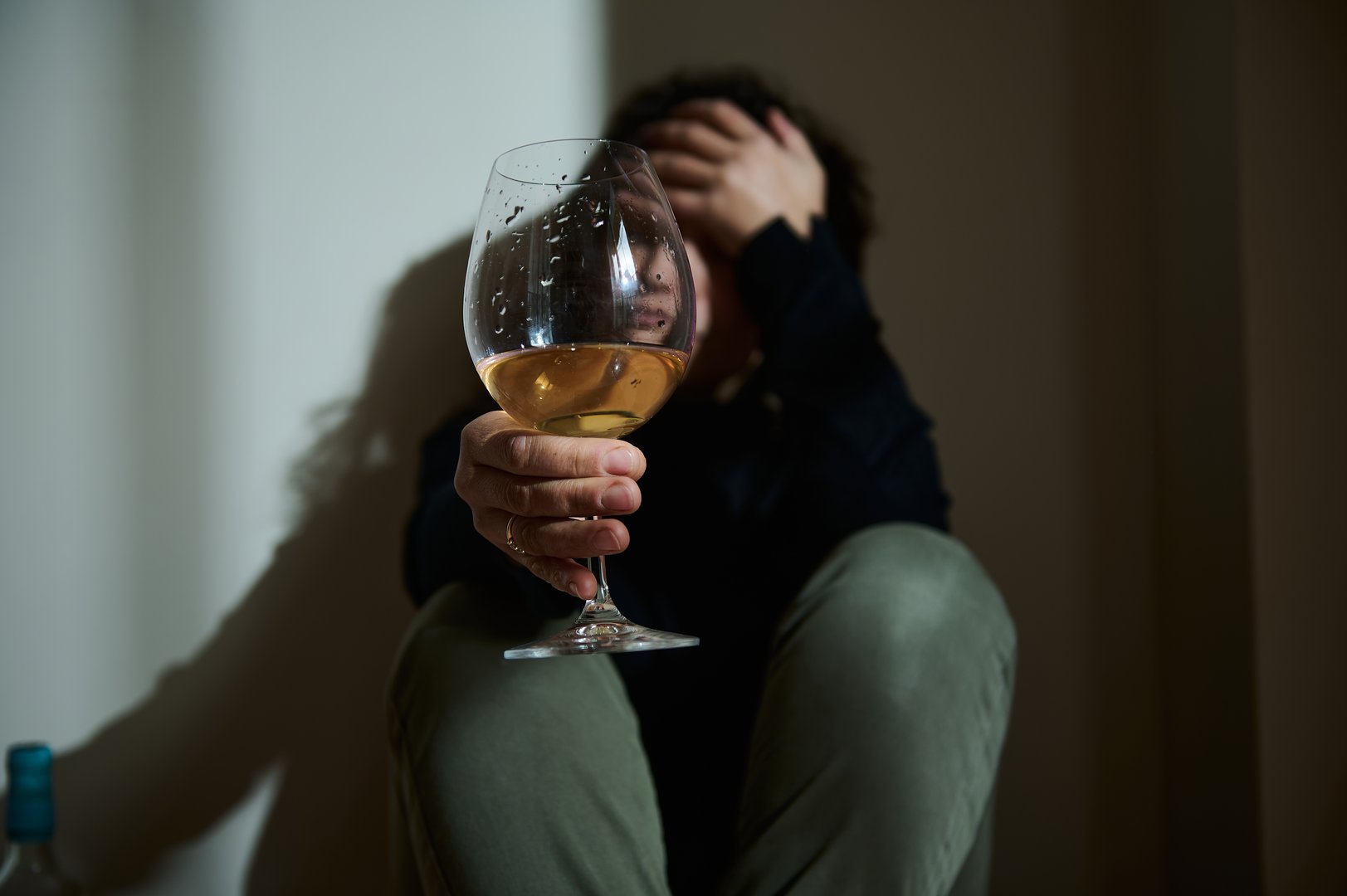 A person seated in low light holds a wine glass, symbolizing emotional distress and self-reflection. The setting conveys themes of internal struggles and moments of personal contemplation.