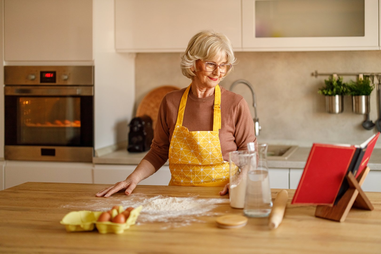 Senior woman with flour and eggs on kitchen island reading recipe book while baking cookies at home