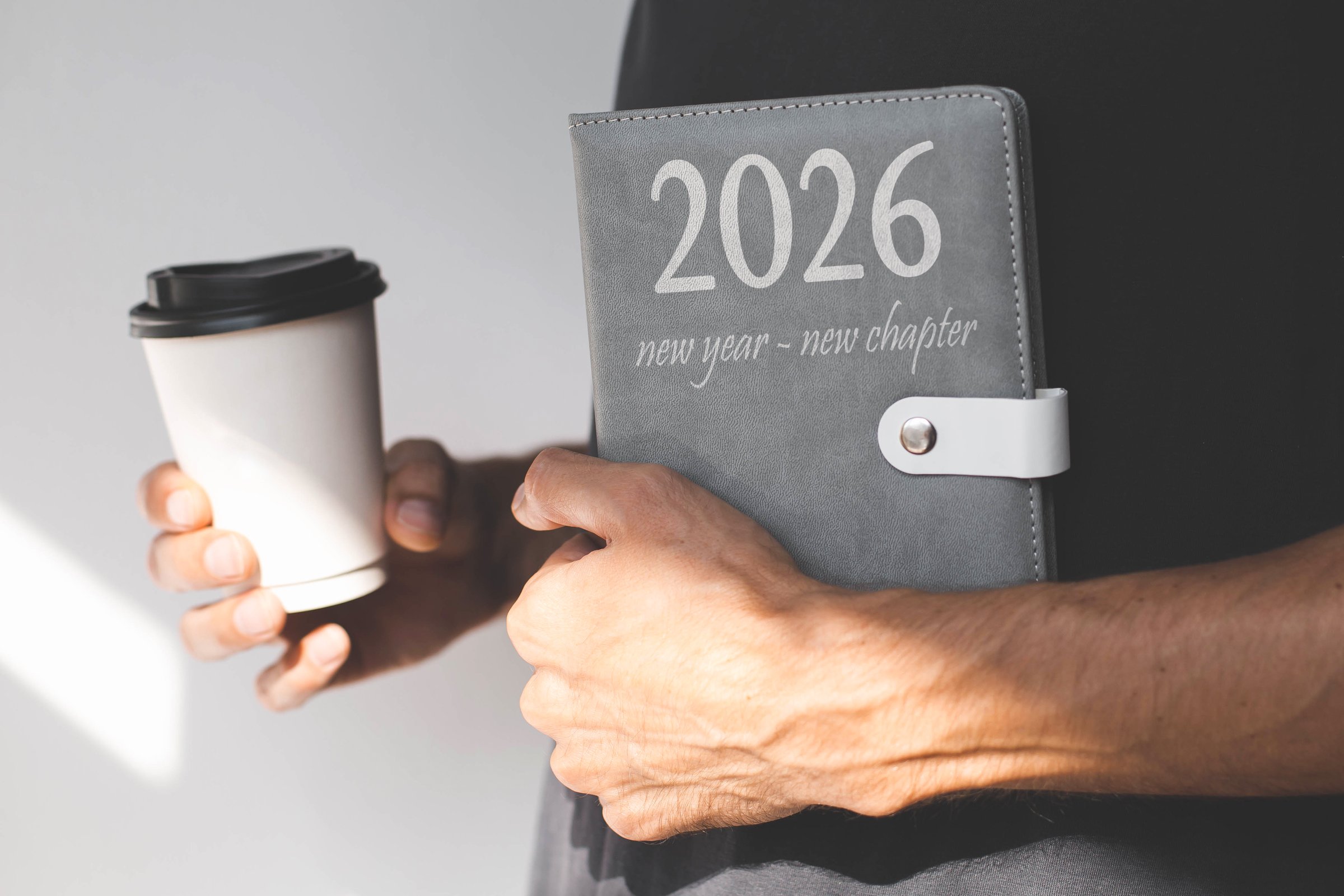 Person holding gray planner with the inscription 2026 new year new chapter takeaway coffee cup, symbolizing motivation, planning, and a fresh start filled with goals, inspiration, and productivity