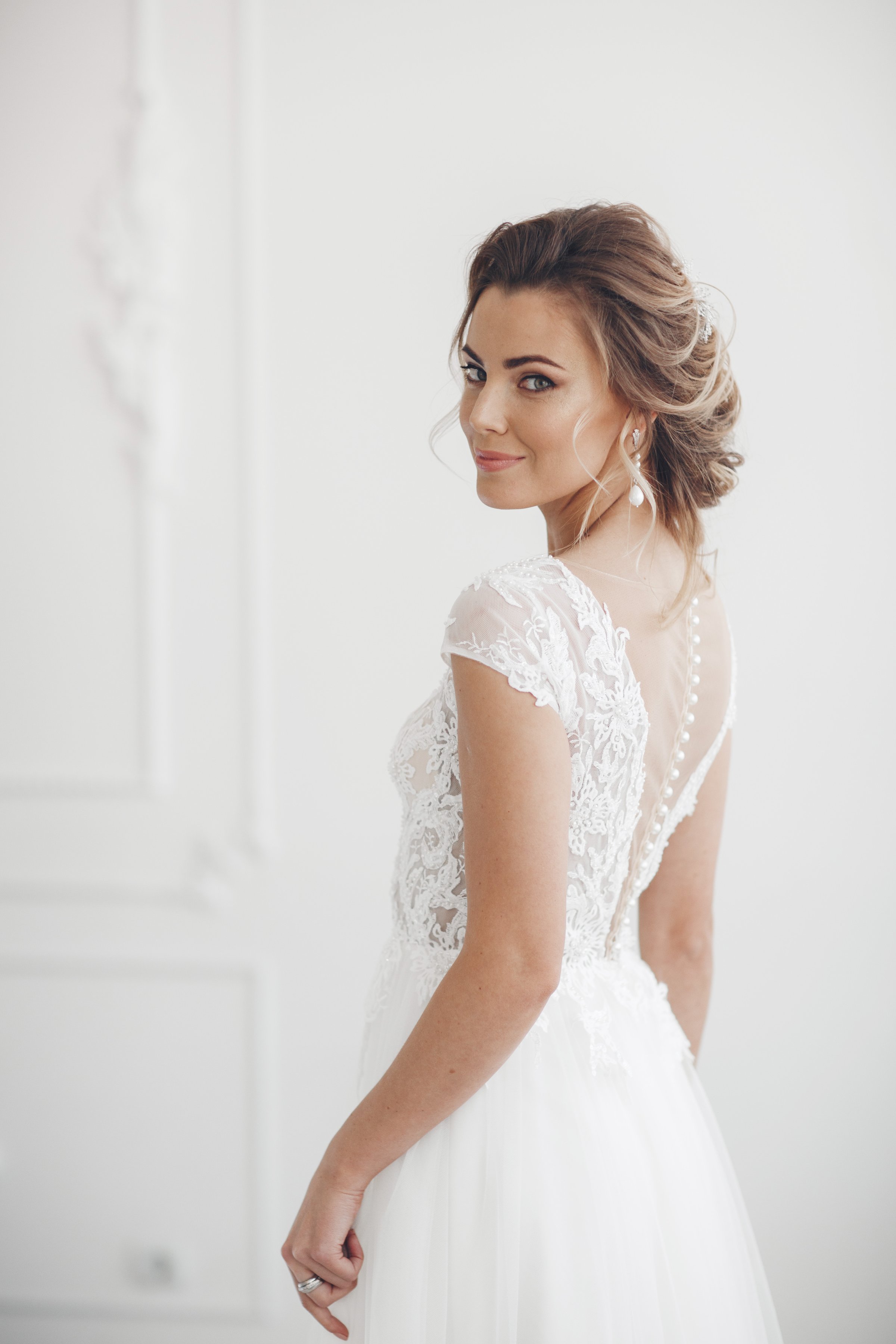 Smiling bride stands alone in a white room. Pretty bride with wedding makeup, fashion jewelry. Beautiful woman posing in white wedding dress. Wedding portrait of happy bride. Bridal fashion model posing in interior. Gorgeous beauty, young bride.