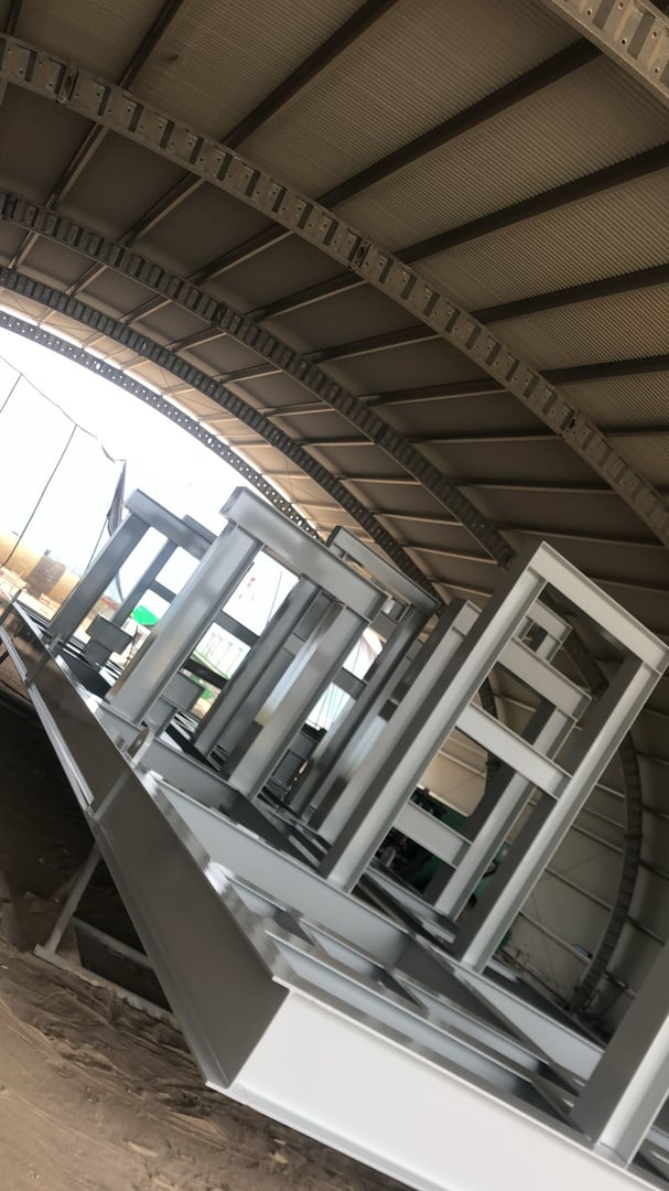 Metal frames under a large, curved industrial structure with beams and a textured ceiling, on a dusty surface.