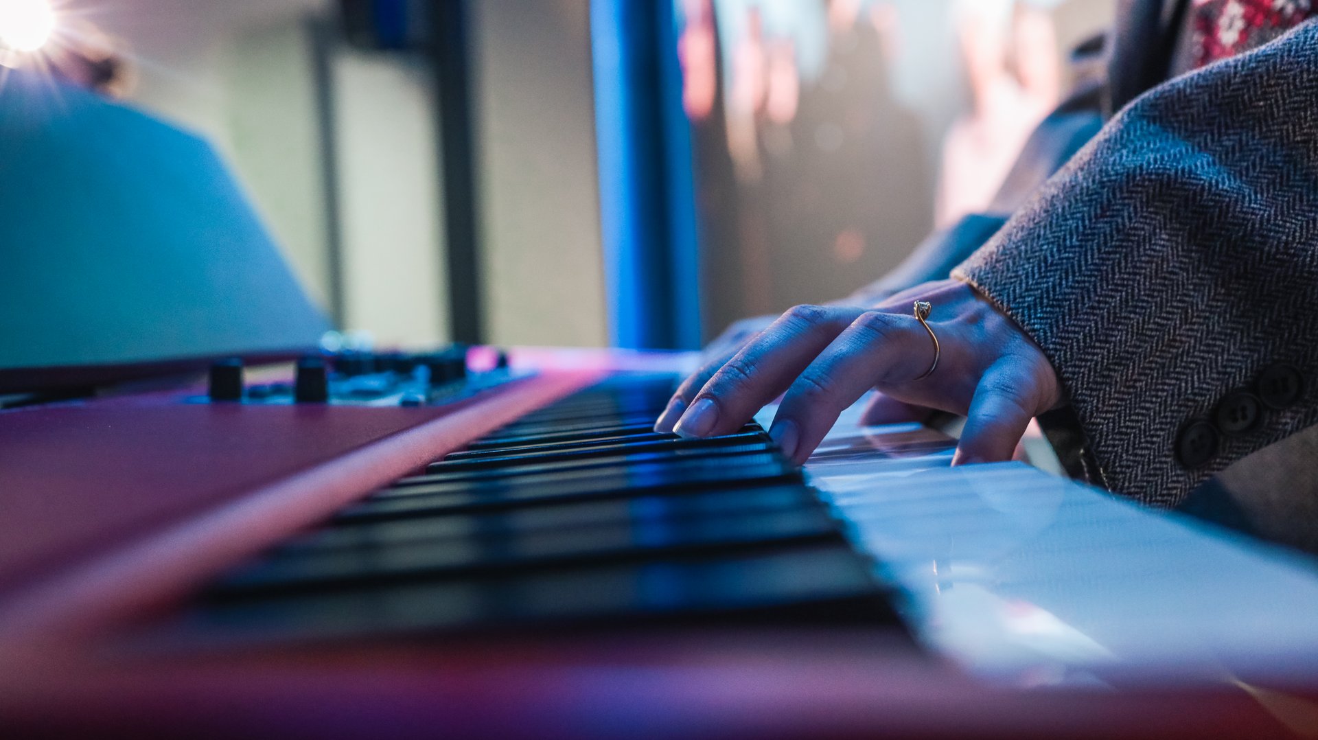 Close-up of hands playing electronic keyboard during live music performance with blurred bassist in background under blue lighting.