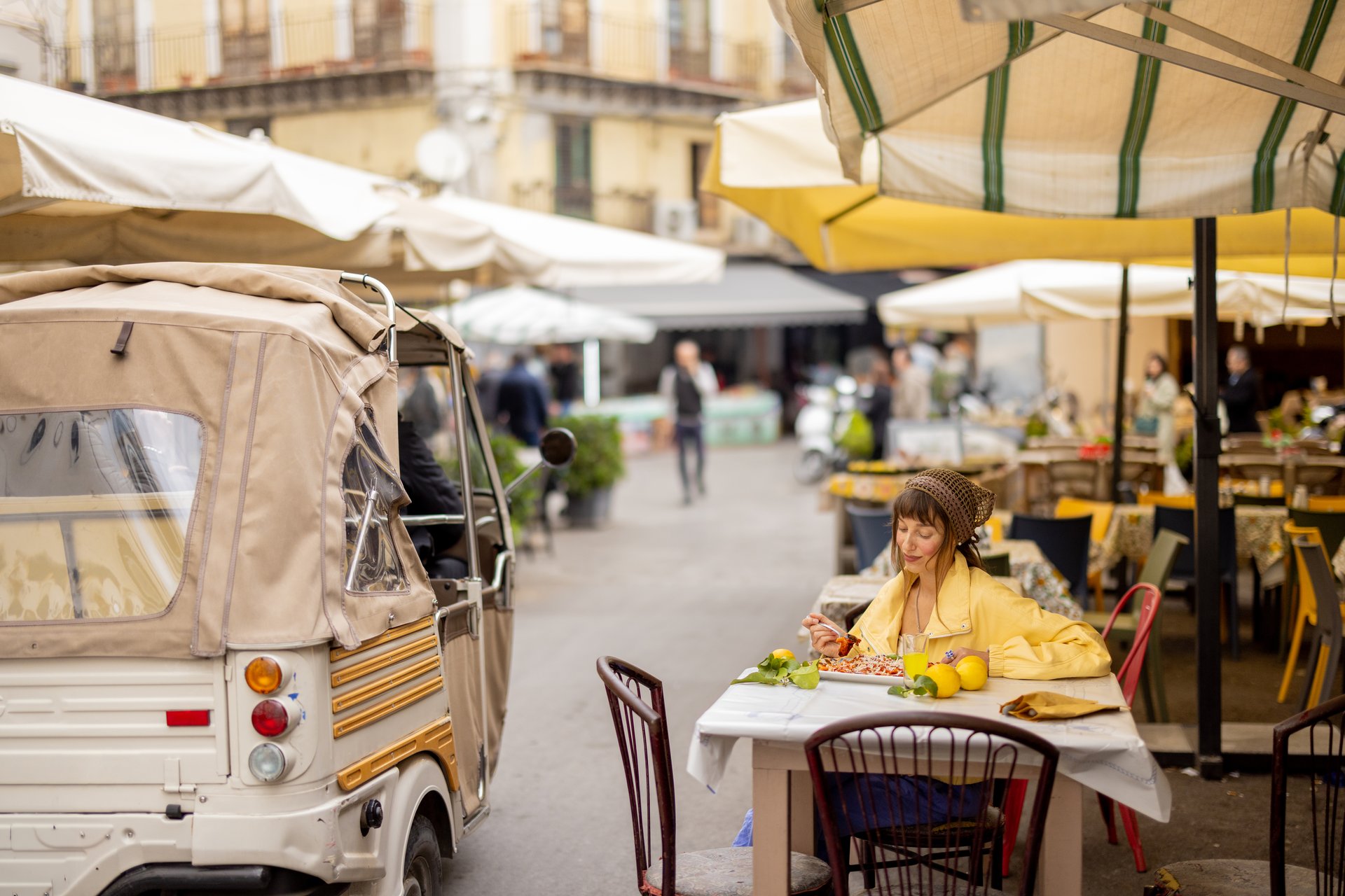 A woman in a yellow jacket enjoys pasta at an outdoor table in a Sicilian street market