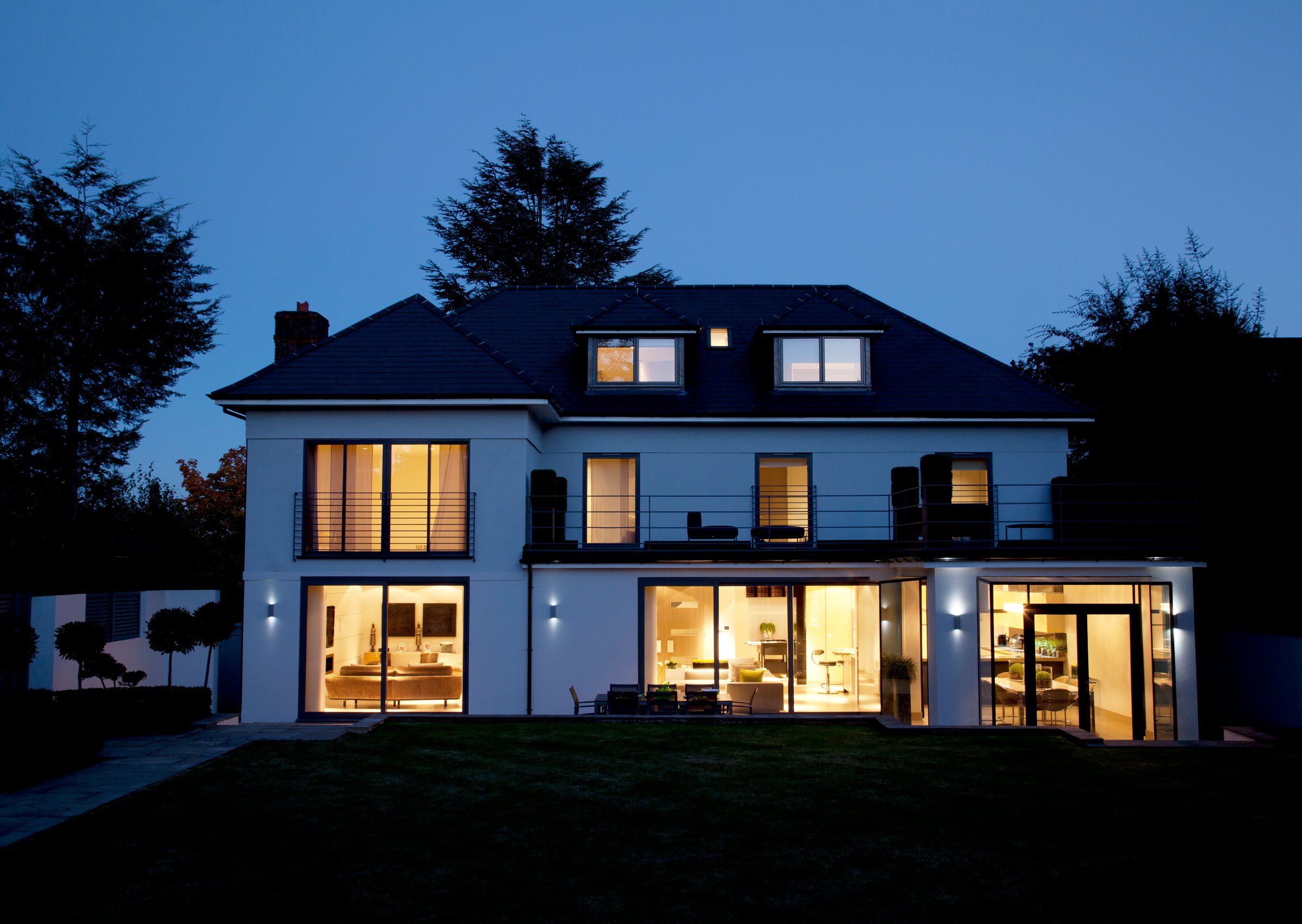 A modern two-story house with lit windows, a patio, and surrounding trees, photographed at dusk.