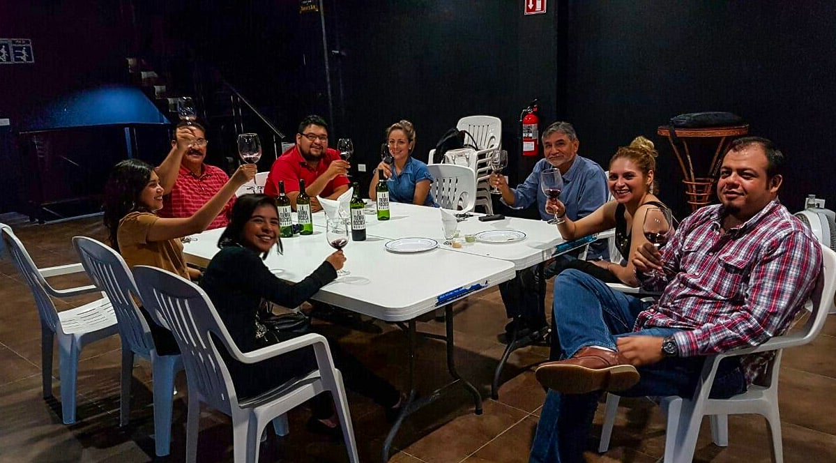Group of people sitting around a table with drinks, raising their glasses in a toast, indoors with plastic chairs.