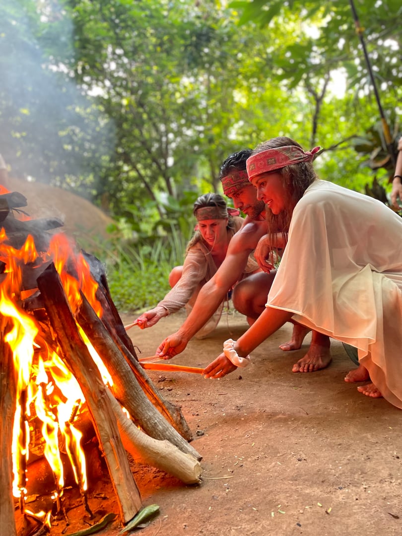 Three people sitting by a campfire in a forest, wearing casual clothes and headbands, engaging with the fire flame.