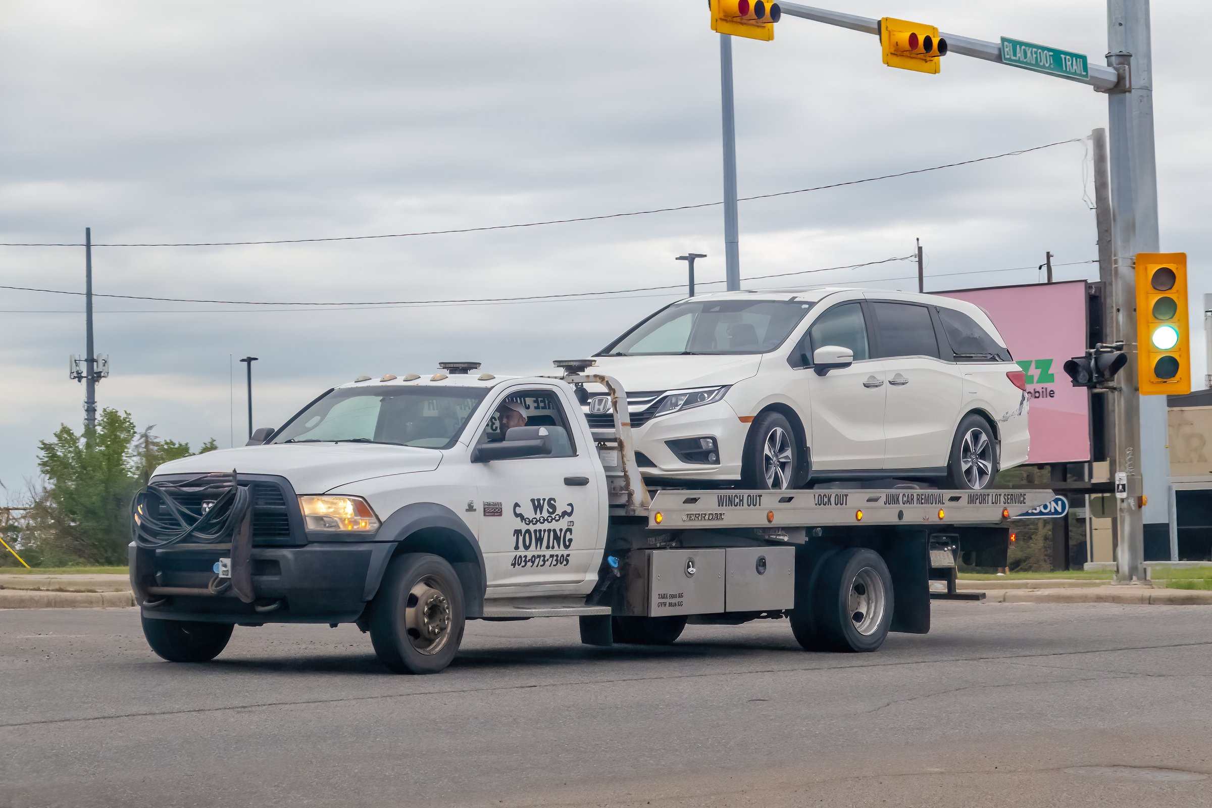 Calgary, Alberta, Canada. Jul 12, 2025. A white W.S. Towing flatbed truck hauling a white minivan on a city street, showcasing roadside assistance and urban transportation.