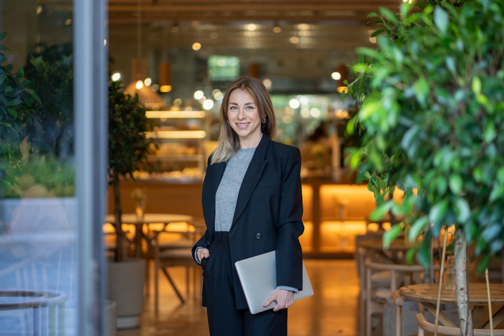 Confident and smiling businesswoman stands with laptop in doorway of brightly lit cafe, looking at camera. Female showing efficiency of internet technology, managing small business and telecommuting