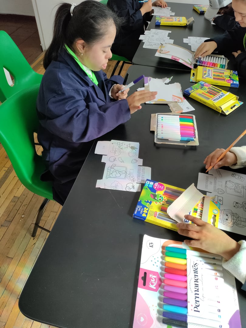 Child coloring on paper with markers at a table, surrounded by coloring supplies, in a classroom setting.
