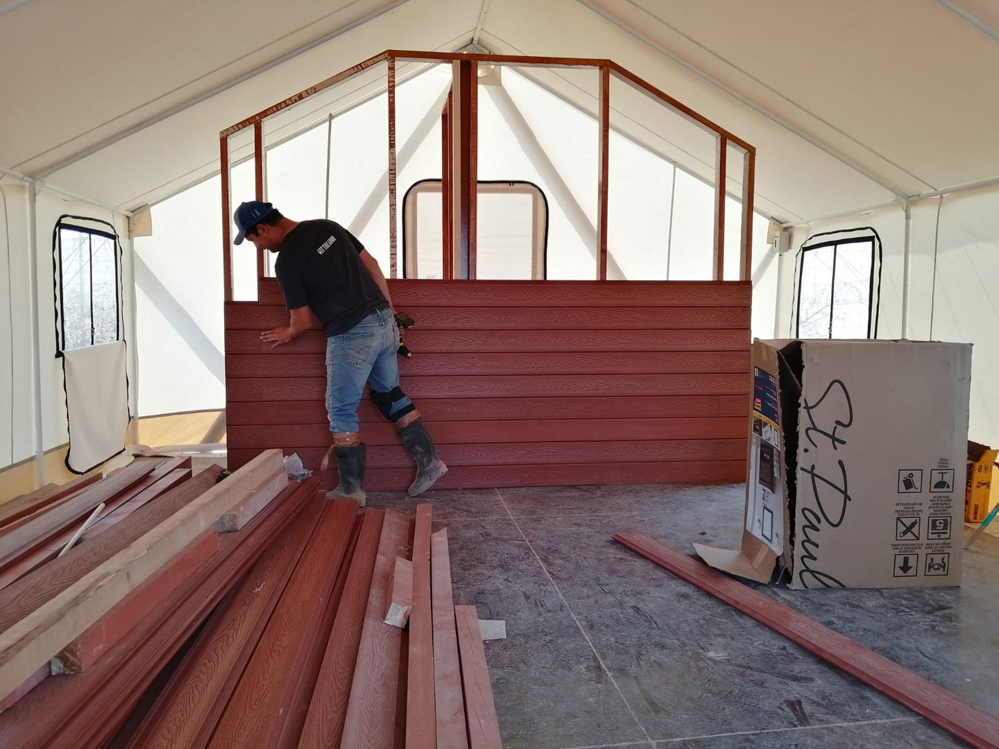 Person installing wood panels inside a partially constructed building frame, surrounded by materials and a large box.