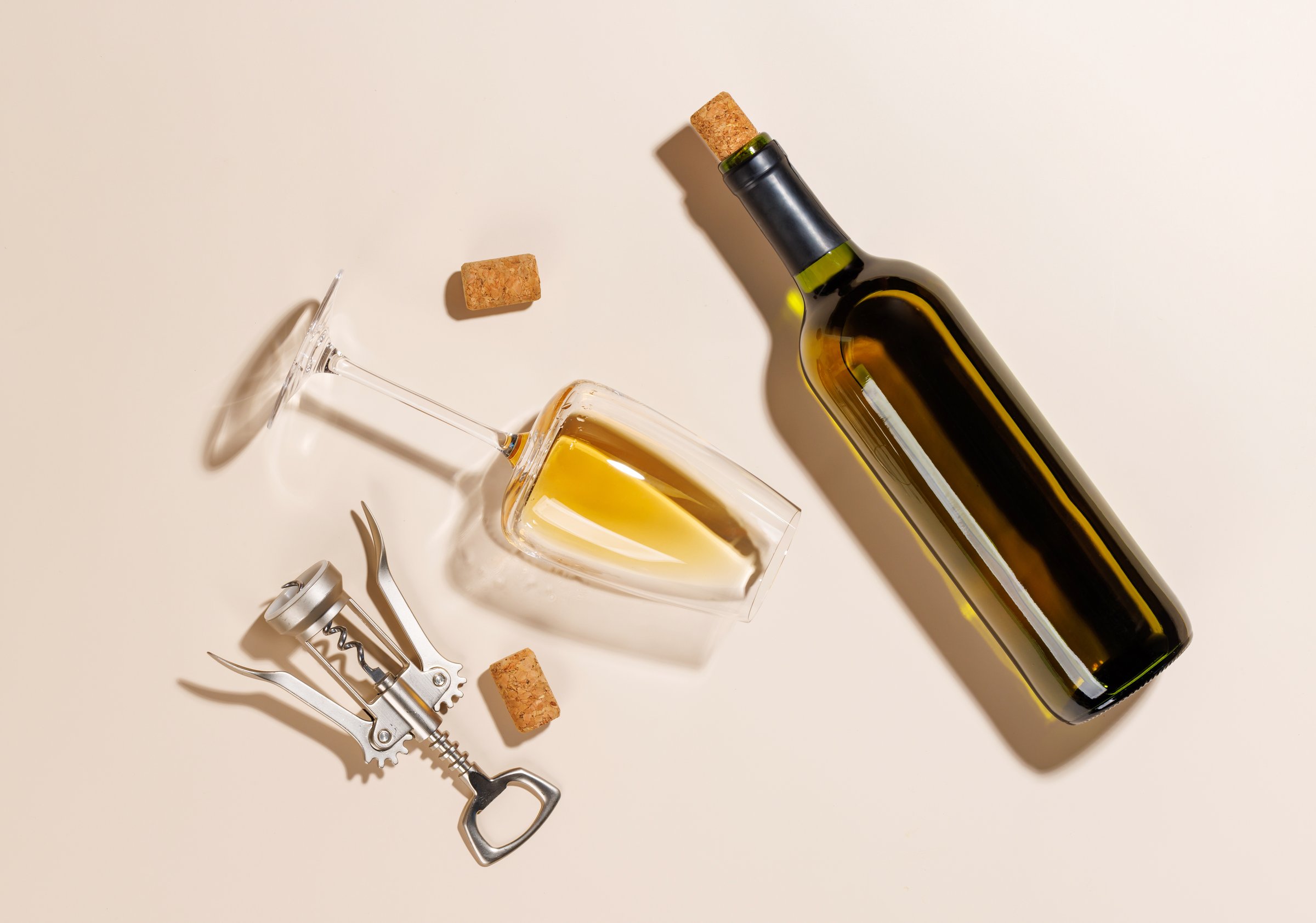 Wine glass, bottle alongside a corkscrew and several corks, all arranged on a bright background. The hard light creates sharp shadows, adding a dramatic effect to the scene, emphasizing the shapes