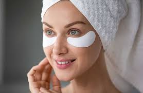 A person with a towel wrapped around their head is wearing under-eye patches and smiling.
