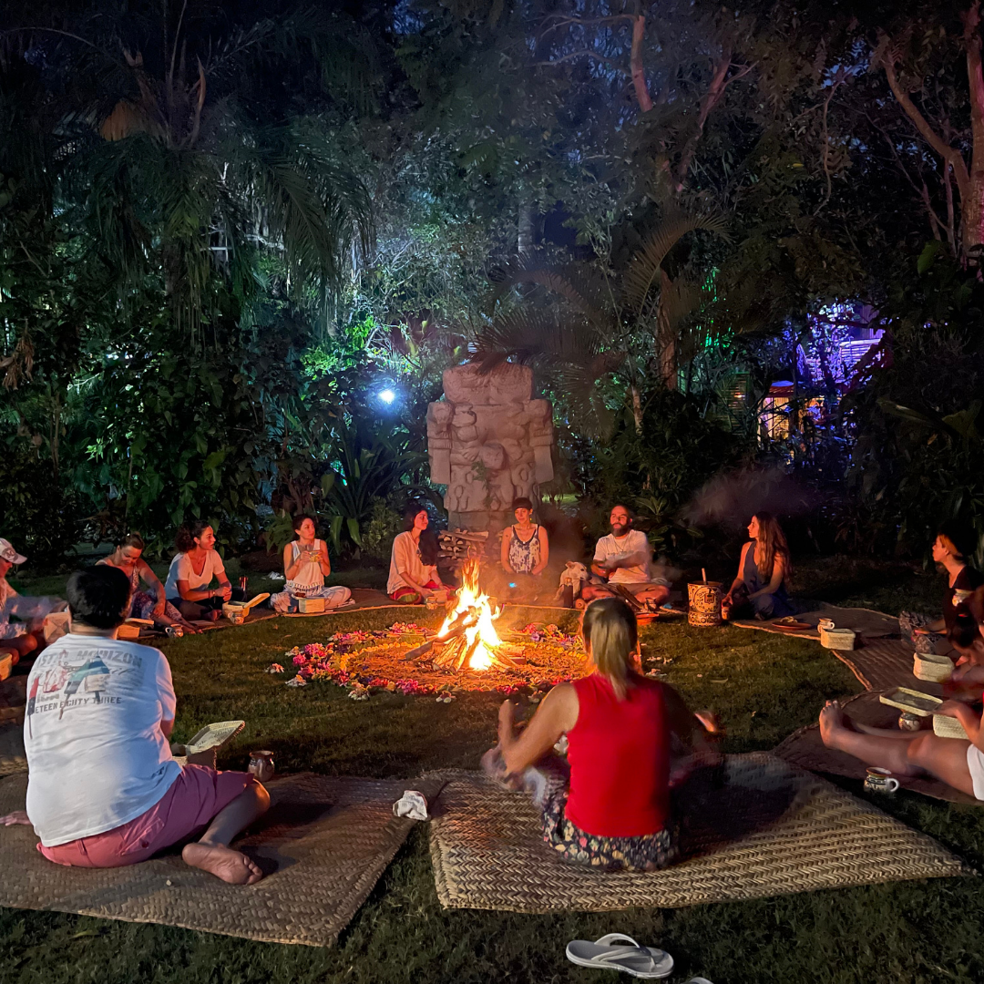 People sitting in a circle around a campfire at night, surrounded by tropical foliage, engaging in a gathering or ceremony.