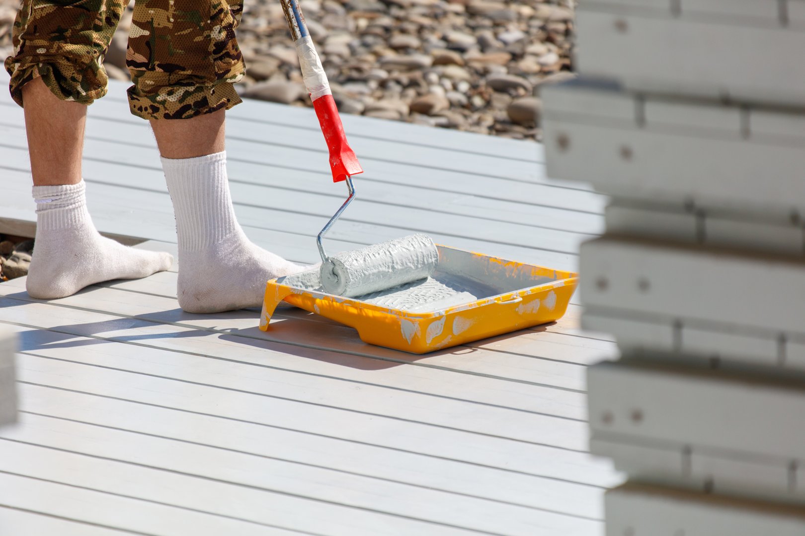 A man is painting a deck with a roller. He is wearing white socks and a camouflage shirt